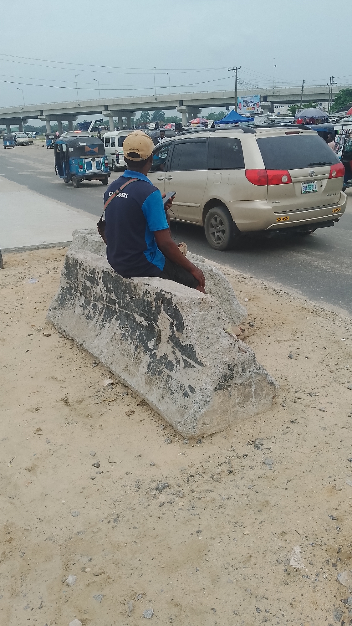 A man in a blue and black shirt and tan cap sits on a large, grey, concrete block in Effurun, Nigeria. He holds a cell phone in his hand. The block is on a sandy area next to a road. A tan Toyota Sienna minivan is partially visible on the road. A blue and black tricycle vehicle is driving away. An overpass bridge is in the background, and an advertising billboard is also visible.