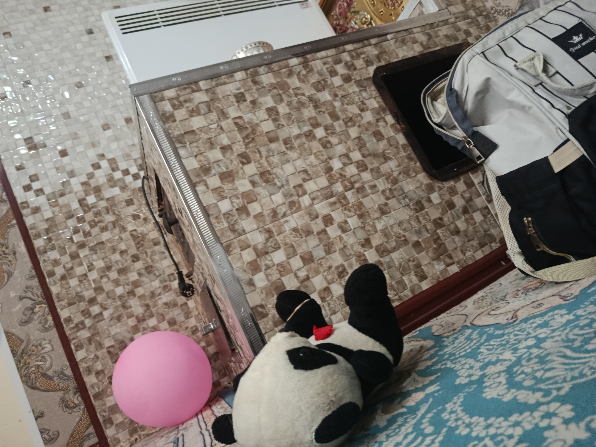 A pink sphere is situated on a patterned carpet next to a plush panda toy. Behind them, a tiled surface leads to a white heating unit. To the right, a backpack rests on the carpet. The scene is captured from a high-angle perspective.