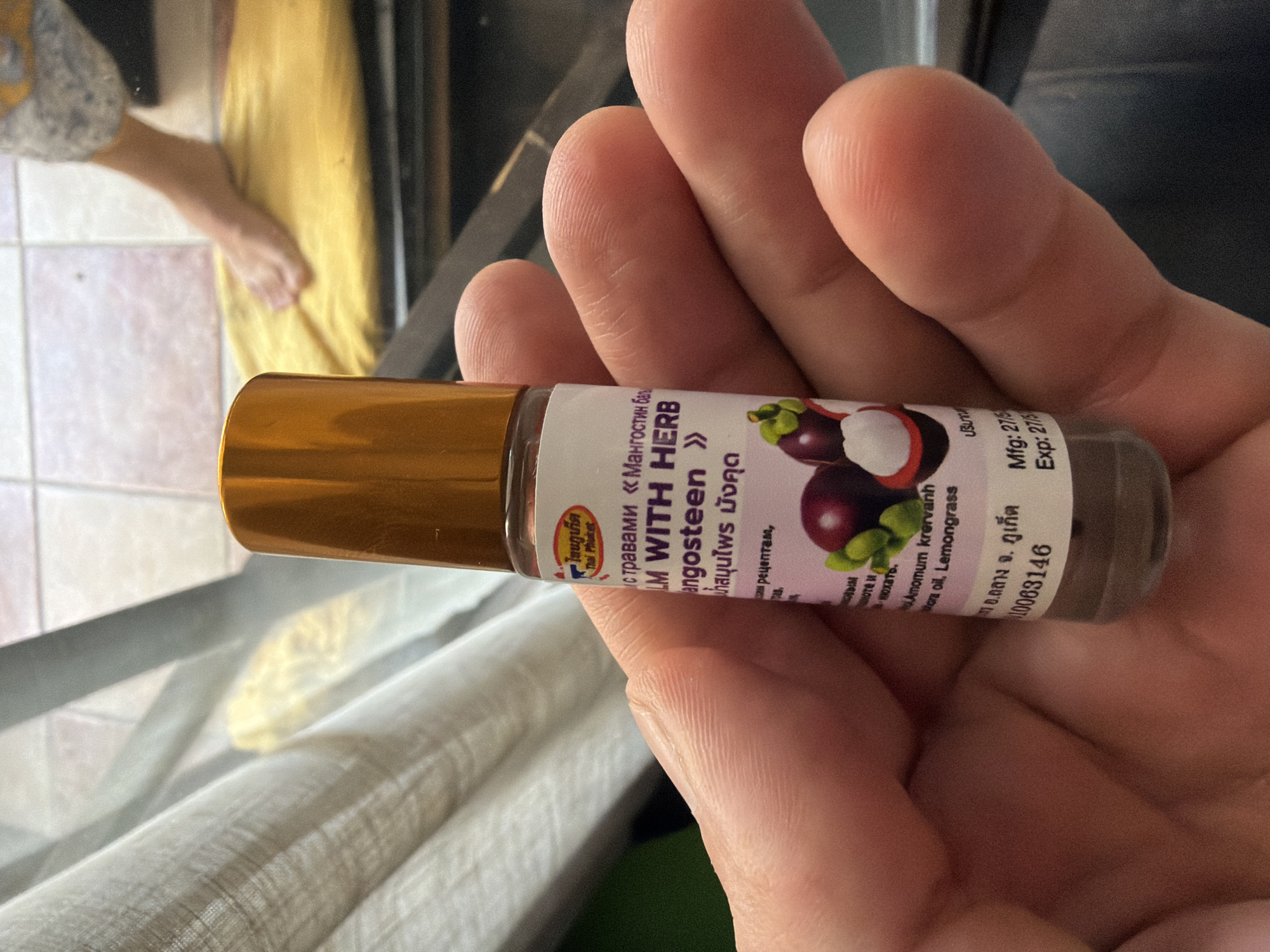 The image shows a close-up of a person's hand holding a small, roll-on bottle of "BALM WITH HERB MANGUSTEEN". The bottle has a gold cap and a label with Thai writing and English text. The label also features an illustration of mangosteen fruit. The background is blurred, showing what appears to be a tiled floor and a person's leg and foot in the distance. There is no visible indication that this image is located in Budva, Montenegro. The focus is entirely on the hand and the product it holds.