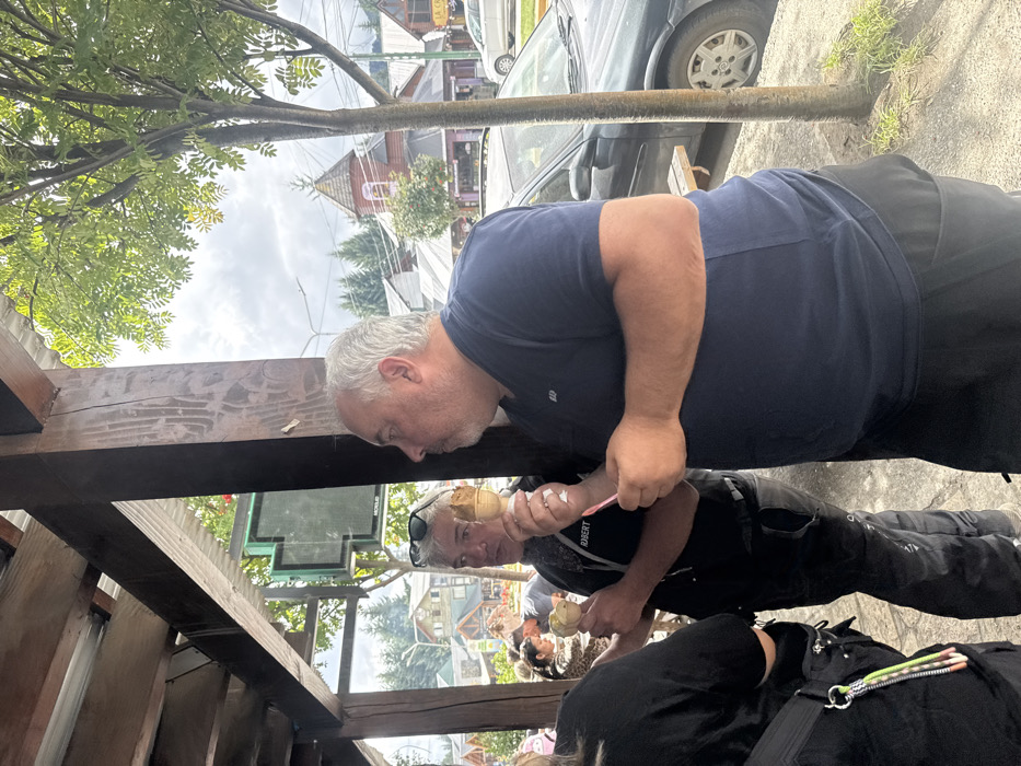 Two men are positioned near a wooden structure, one behind the other. The man closer to the foreground is older, with gray hair, wearing a dark blue shirt and black pants. He is looking down at an ice cream cone held by the man in front. The man in front, also older with gray hair and sunglasses, is wearing a black shirt with white writing. The background shows a street scene with buildings, trees, and a car. The photograph is taken at an unusual angle.