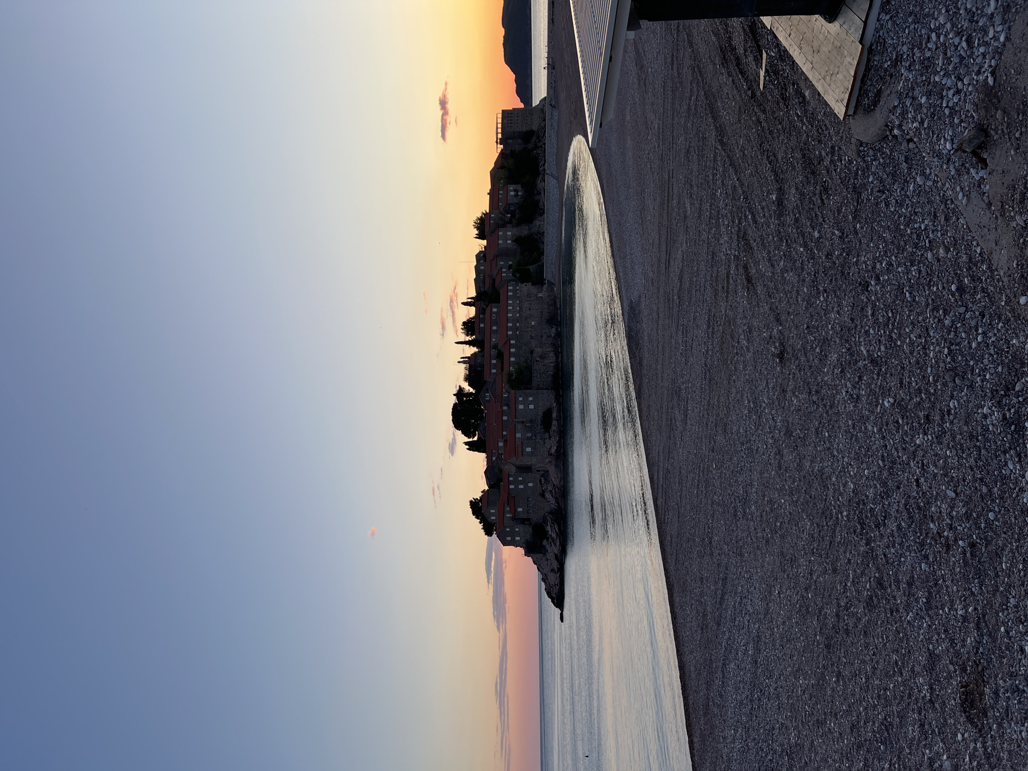 The image, correctly oriented in landscape format, presents a picturesque coastal scene of Sveti Stefan, Montenegro, captured during either sunrise or sunset. The sky dominates the upper portion, displaying a gentle gradient from soft blue and light purple on the left to warm, vivid orange and yellow on the right, characteristic of the golden hour. A few small, ethereal clouds are scattered across the sky, echoing its colors.
The central subject is the distinctive fortified island settlement of Sveti Stefan, located towards the left-center. It is densely packed with stone buildings, each featuring traditional red-tiled roofs, nestled among dark green foliage. The island extends into the calm, reflective waters of the Adriatic Sea, which mirrors the subtle hues of the sky.
To the right of the island, a wide pebble beach stretches towards the foreground, with lighter pebbles forming a path along the water's edge. The beach transitions to a darker, coarser-grained surface closer to the viewer. On the far right, a modern wooden and concrete walkway or pier structure is partially visible, suggesting nearby amenities. In the far distance, the silhouette of mountains provides a dramatic backdrop against the colorful horizon.
The scene conveys a sense of tranquility and natural beauty, emphasizing the historic charm of Sveti Stefan at a serene time of day. No human activity or visible text is discernible in the image.