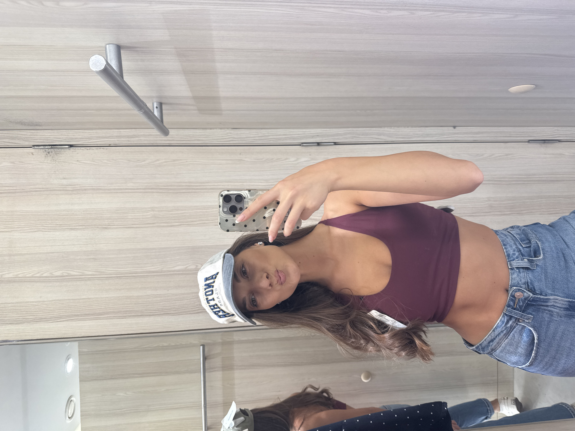A young woman is taking a selfie in what appears to be a fitting room. She is wearing a maroon crop top, blue jeans, and a baseball cap with the word "GEORGETOWN" on it. She is holding a smartphone with a spotted case in her right hand. The fitting room has light-colored wooden paneling. In the mirror, a reflection of another person in navy blue, jeans, and white shoes can be seen. The location cannot be specifically determined as being in Burgas, Bulgaria, from the given information.