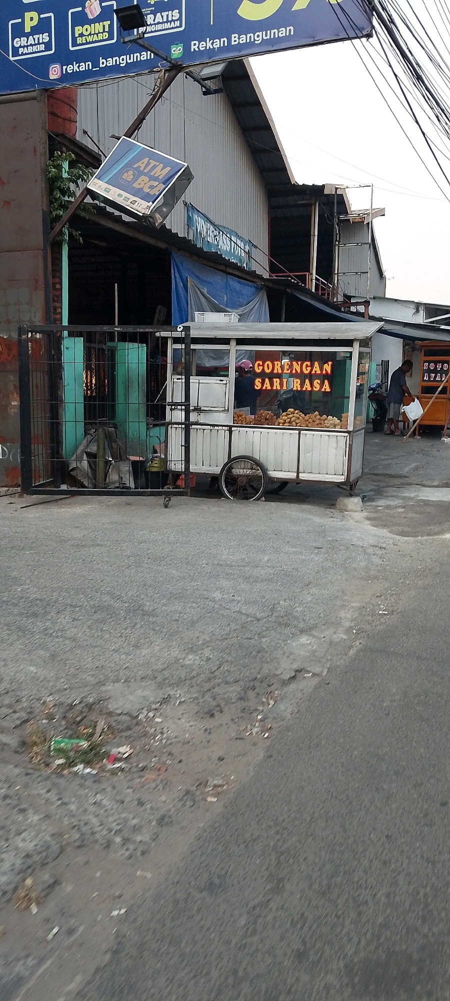 The image shows a street scene in Bekasi, Indonesia, featuring a food cart. The food cart is white with a glass display case filled with fried foods. Neon orange text on the cart reads "GORENGAN SARI RASA". A person in a pink hat is visible inside the cart. Next to the cart, a man in a blue shirt is interacting with another vendor's cart, which displays "SOTO AYAM". A black metal gate is to the left of the food cart, obscuring some of the building behind it. An ATM BCA sign is tilted precariously on a pole above the buildings. The ground is paved with asphalt, showing signs of wear and tear with some debris scattered on it. The background includes buildings with corrugated metal siding and tarpaulin coverings. The overall scene depicts a commercial area with food vendors.