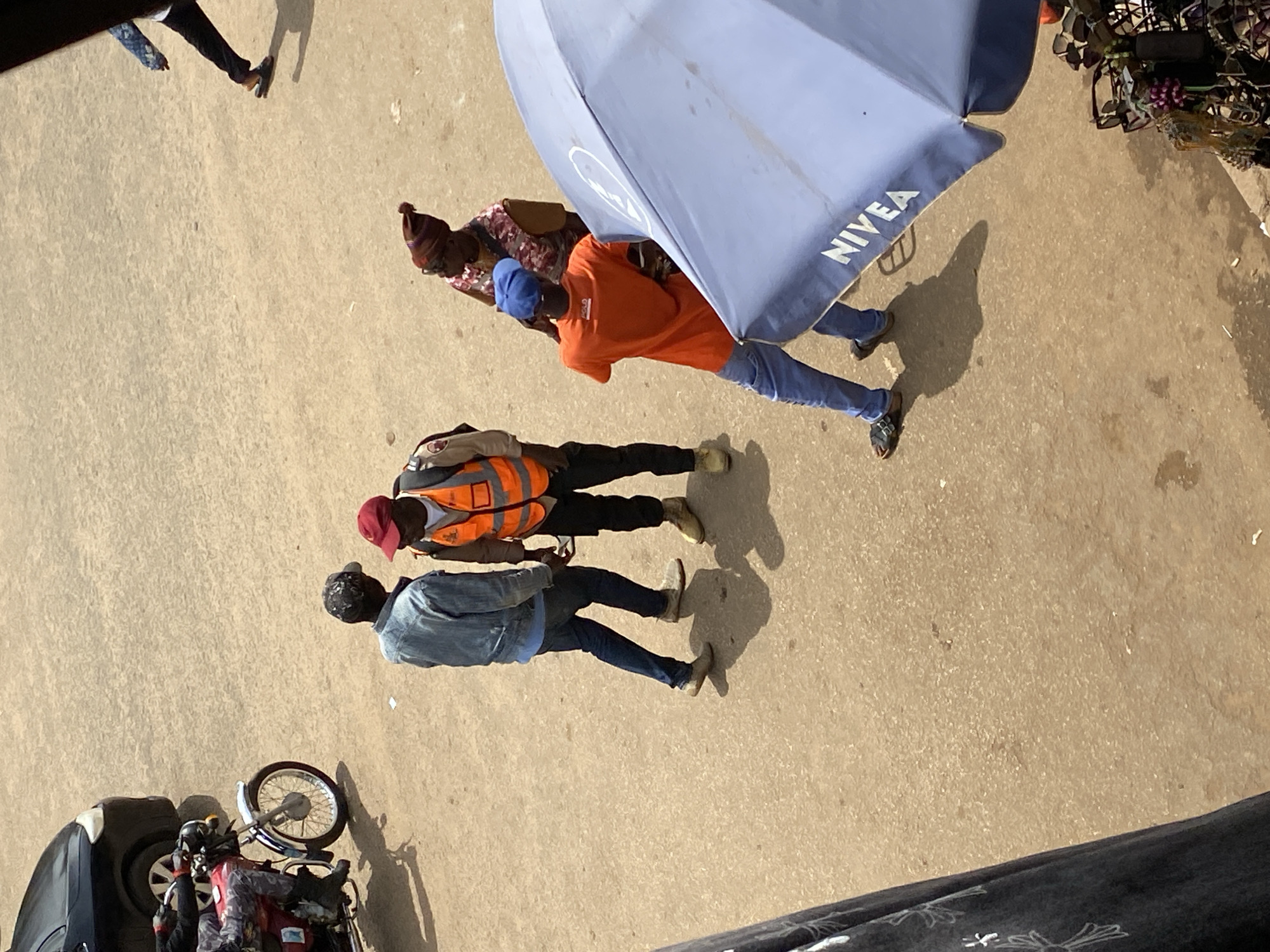 The image depicts an outdoor scene on a street in Bukuru, Nigeria, characterized by a dusty, sandy road surface and bright sunlight casting distinct shadows. In the mid-ground, four individuals are present. Two men, one wearing a denim jacket and the other an orange safety vest over a white shirt, are conversing. To their right, a woman in a patterned top and a beanie hat looks upwards. Further to the right, a man in an orange t-shirt and blue jeans is talking, his back mostly to the camera. A motorcycle with a rider is parked in the background on the left. The foreground is partially occupied by a large blue umbrella with the brand "NIVEA" visible, positioned to the right and angled towards the viewer. To the far left, a dark fabric with a white floral pattern is visible, suggesting a stall or structure. The overall atmosphere suggests a bustling urban environment.