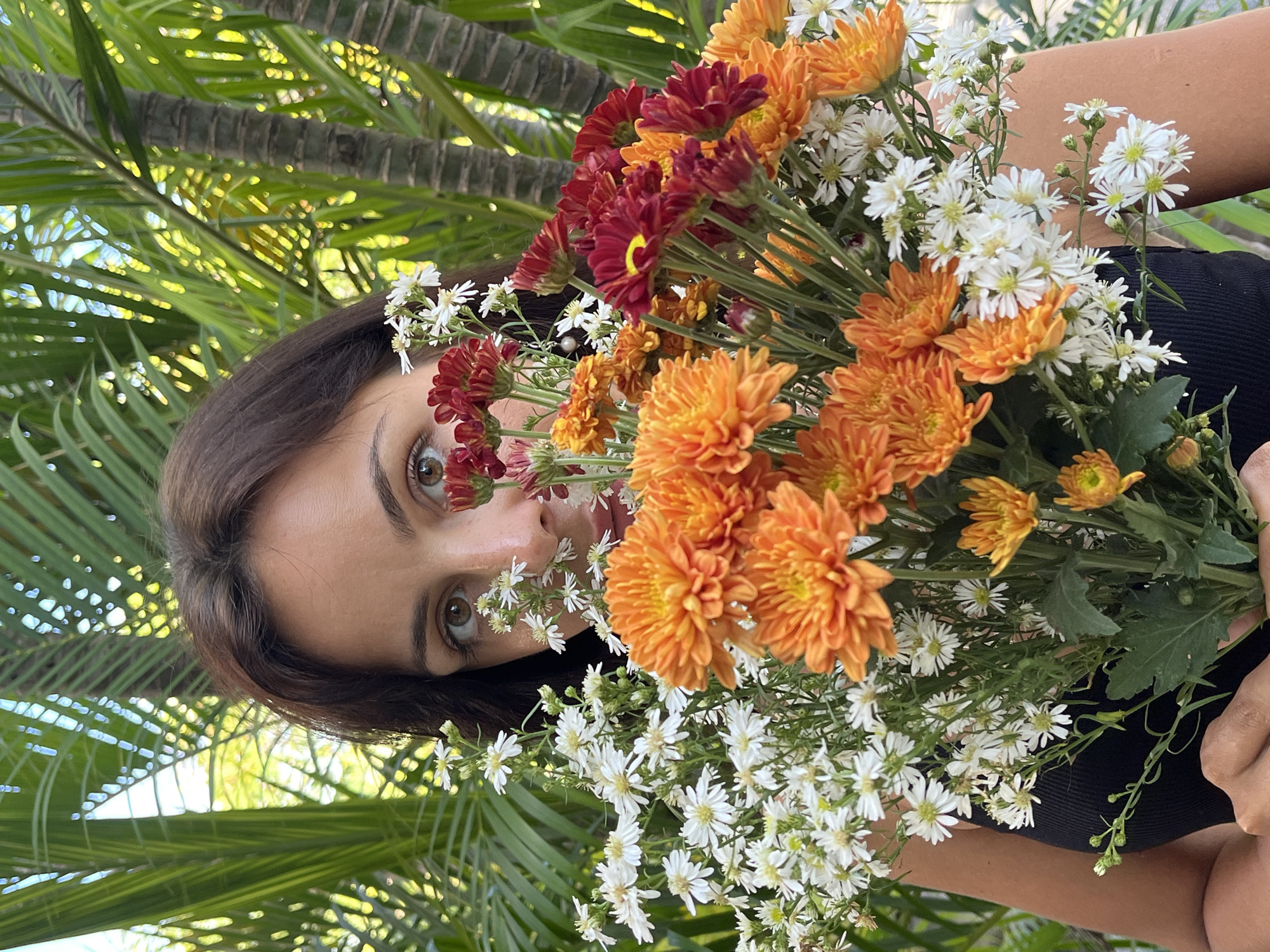 A portrait-style photograph features a woman holding a large, vibrant bouquet of flowers that partially obscures her face. The woman, with dark brown hair and a tanned complexion, gazes directly at the viewer with wide, brown eyes that peek through the blooms. She is wearing a black top or dress, with her arms visible as she holds the bouquet.
The bouquet is rich in color and texture, composed primarily of bright orange and deep red chrysanthemum-like flowers, interspersed with numerous smaller, delicate white daisy-like blooms. The flowers are arranged densely, creating a natural frame around the woman's visible features.
In the background, lush green palm trees and dense foliage are clearly visible, indicating an outdoor, tropical setting. The lighting is bright and natural, suggesting it is daytime. The overall scene conveys a fresh and natural aesthetic. No visible text is present in the image. The location context provided is Kuta Selatan, Indonesia.