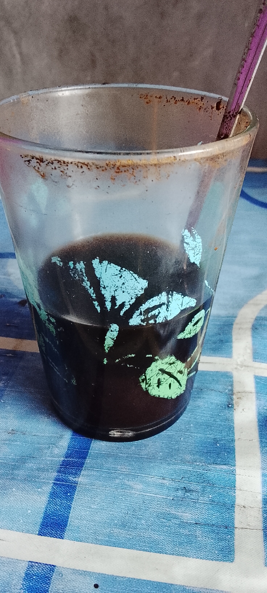 The image shows a close-up view of a glass of dark liquid, likely coffee, with a spoon inserted. The glass has a faded pattern of blue and green motifs. The rim of the glass is stained with brown residue, and there are also some smudges on the exterior. The glass sits on a blue and white checkered surface, possibly a tablecloth or placemat. The background is out of focus and appears to be a textured wall. No people are visible in the image. The location is specified as Padang Dalom, Indonesia.