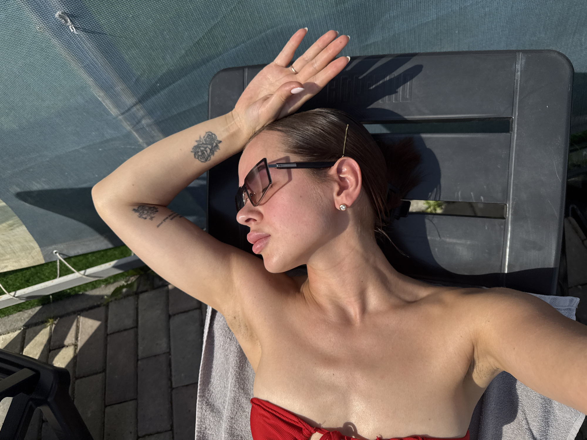 A woman is lying on a black plastic outdoor chair under a partially transparent blue canopy, possibly in Гамово, Russia. She is wearing sunglasses and a red top. Her left arm is raised with her hand above her head. She has two tattoos on her left arm. A white towel is underneath her. The ground is paved with gray brick.