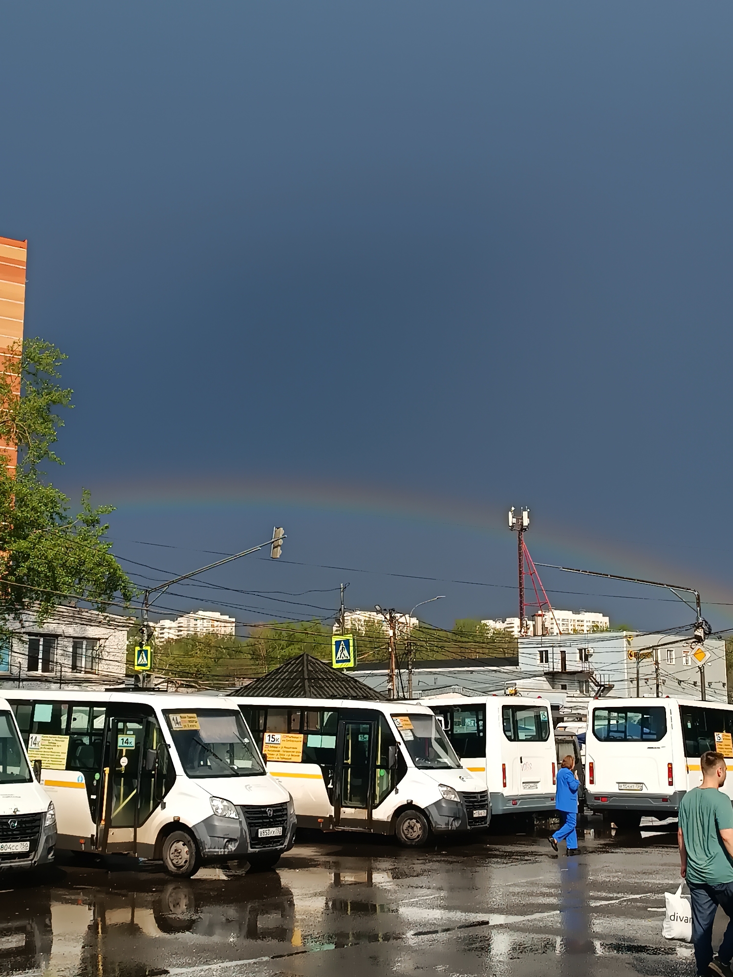 The image shows a street scene in Lyubertsy, Russia after a rain shower, with a rainbow visible in the background. In the foreground, a row of white vans, possibly minibuses for public transportation, are parked on a wet asphalt surface, their reflections mirrored in the puddles. One of the vans has an open door. A woman in a blue jumpsuit walks between the vans, and a man in a green shirt and jeans carries a white bag labeled "divan" as he walks away from the camera. Overhead are power lines and traffic lights. Behind the vans, there are buildings of different heights and architectural styles, including an orange brick building visible on the left edge. A communications tower is visible in the mid-ground, intersecting the rainbow. The sky is dark, suggesting the storm has recently passed.