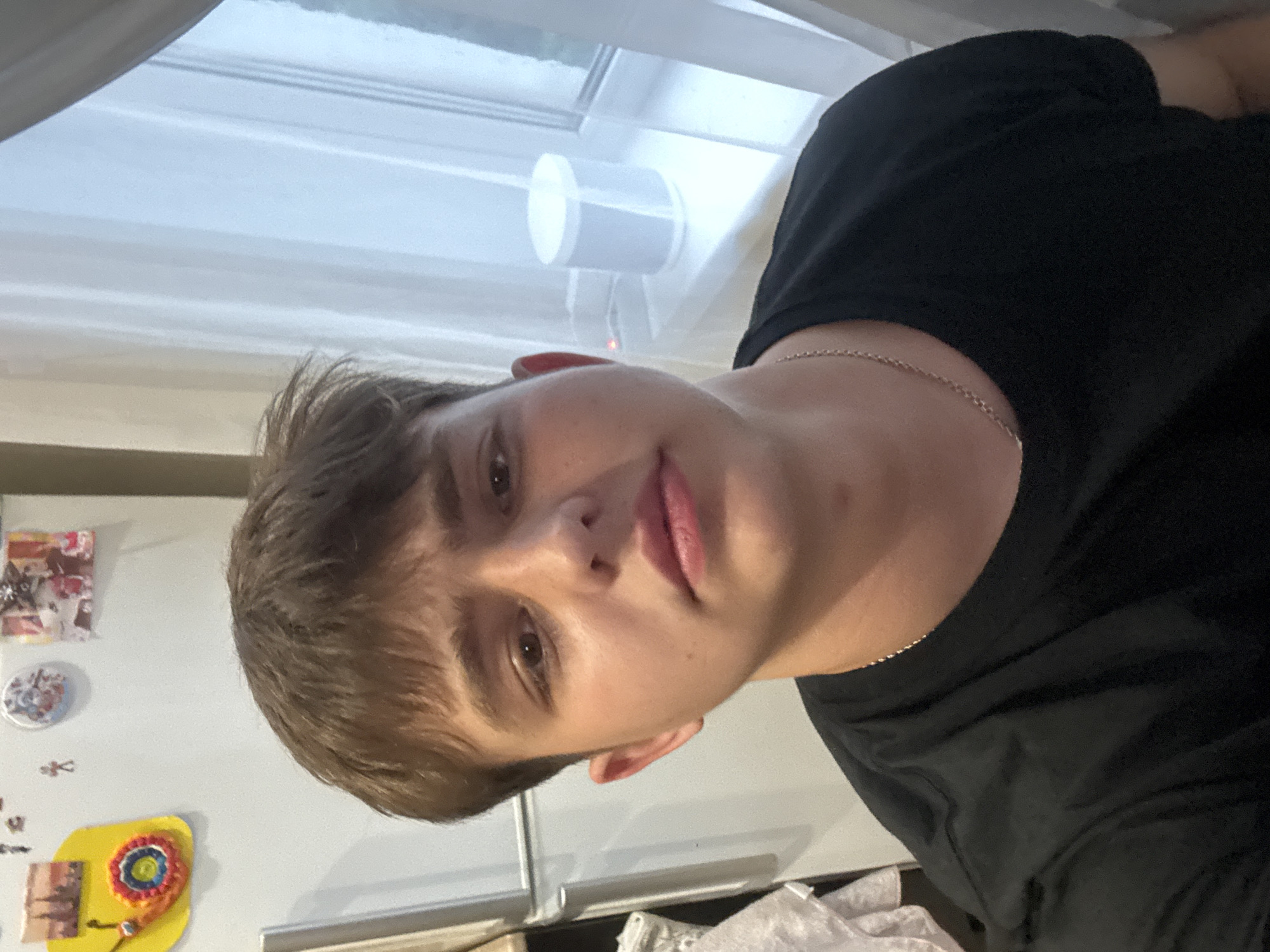 A young male with brown, tousled hair is looking directly at the camera, taking a selfie. He is wearing a black t-shirt and a silver chain necklace. The background features a white refrigerator adorned with various magnets and pictures. To the right, a window with sheer white curtains is visible, and on the windowsill, a white cylindrical object, possibly a speaker, rests. The setting appears to be an indoor environment, likely a kitchen or living area. There is no indication of the city of Birobidzhan, Russia.