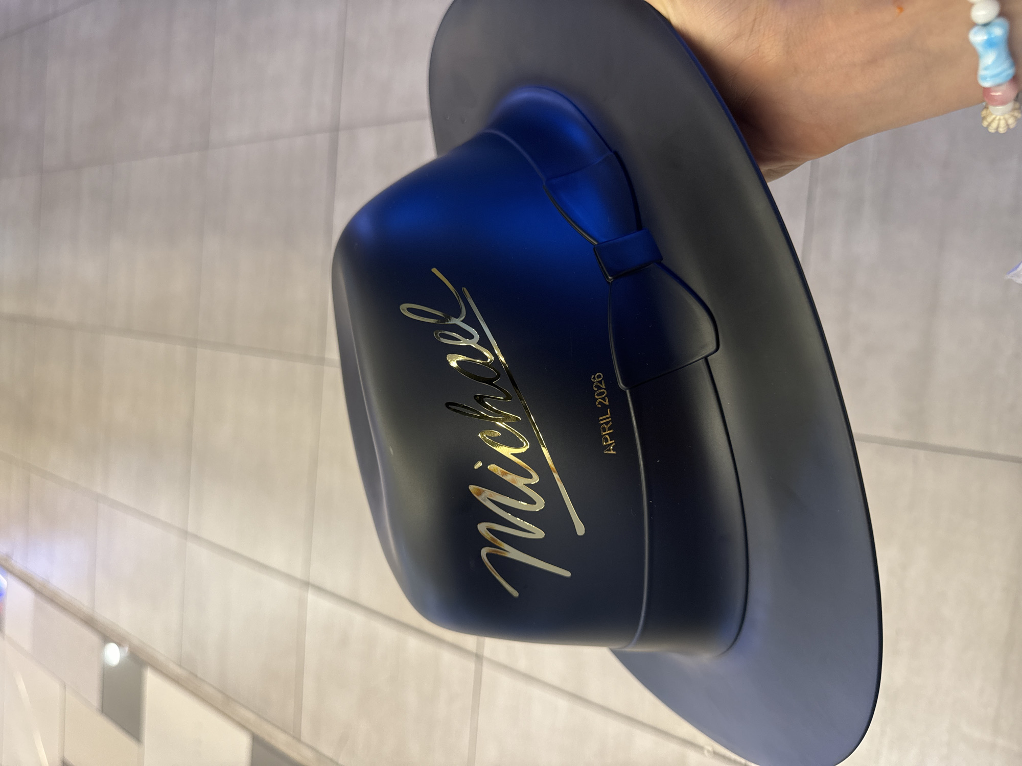 A person is holding a dark blue hat. The hat has "Michael" written in gold cursive on one side and "APRIL 2026" written in gold capital letters on the other side, near the bow. The person is wearing a beaded bracelet with pastel colors. The background appears to be a tiled wall, possibly in a bathroom or kitchen. The location is unknown; there is no information to suggest it is in Ho Chi Minh City, Vietnam.
