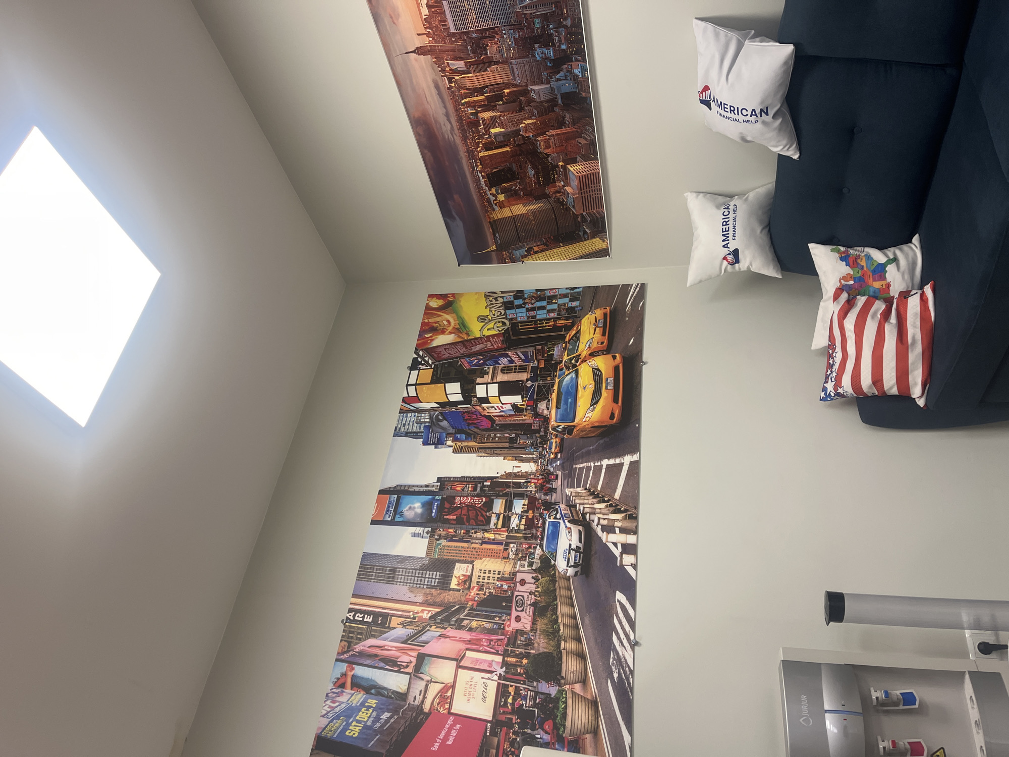The image is an interior shot of a room with a modern aesthetic. Dominating the wall are two large prints: one depicting a cityscape at sunset, and another showcasing a vibrant street scene with yellow taxis. A dark blue couch sits against the wall with several throw pillows, some branded with "American Financial Help", others with colorful designs and the American flag. The wall behind the couch is a neutral gray. A water cooler with a silver and gray finish is positioned near the wall. A skylight provides illumination. The scene does not seem to be in Yerevan, Armenia, but rather New York City, judging by the subject matter of the prints and pillows.