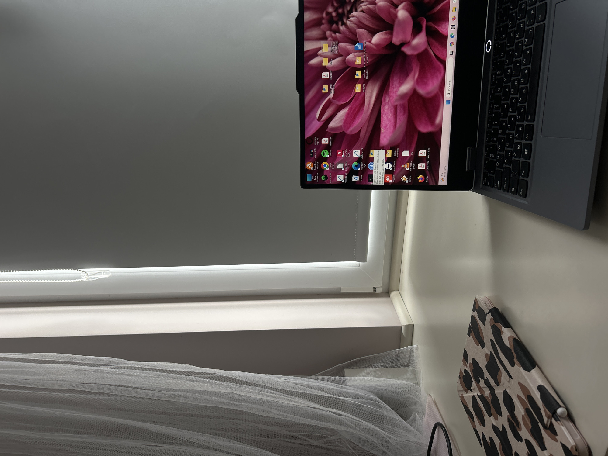 The image shows a laptop with a pink floral wallpaper open on a desk next to a window. The laptop screen displays various icons and files. A leopard-print tablet is placed on the desk to the left of the laptop. The window has sheer white curtains and a white roller blind. The overall scene is an indoor workspace with natural light coming from the window. There is no indication of the location being Plovdiv, Bulgaria.