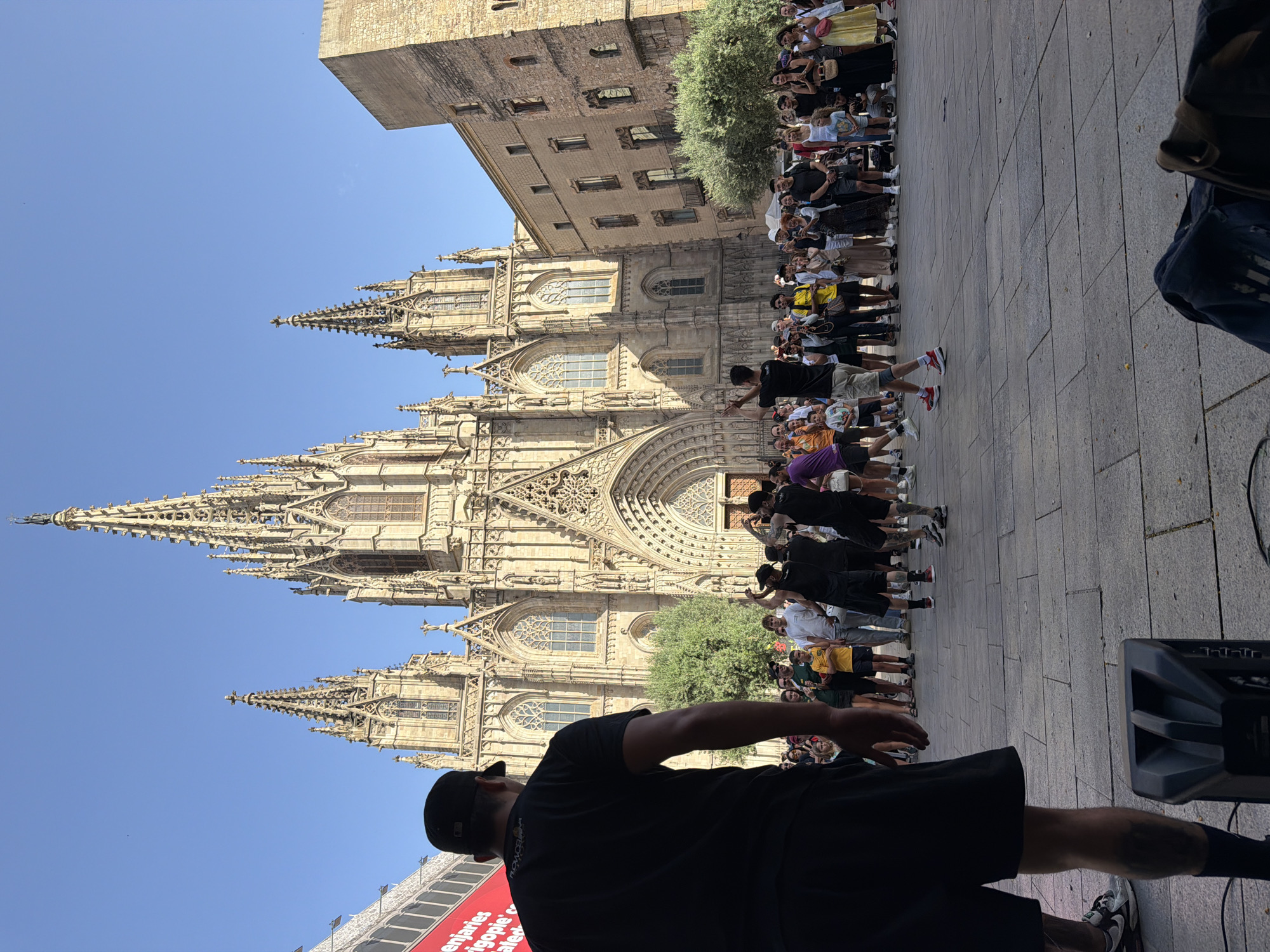 The image depicts an outdoor performance in front of the Barcelona Cathedral, also known as the Cathedral of the Holy Cross and Saint Eulalia, located in the Gothic Quarter of Barcelona, Spain. The cathedral's imposing Gothic facade, complete with spires and intricate stonework, dominates the background. A crowd of people has gathered to watch a group of performers in the foreground.
In the immediate foreground, a person wearing a black t-shirt and black shorts stands with their back to the camera, facing the performers and the crowd. This individual appears to be part of the performance setup, possibly an organizer or DJ, as a black speaker is visible at their feet.
The central action involves a group of male performers on the paved plaza in front of the cathedral. Three men are in the center, one wearing a black t-shirt and shorts, another in a white t-shirt and dark shorts, and a third in a black t-shirt and dark shorts. They appear to be engaged in some form of street performance, perhaps dancing or a martial arts demonstration, with dynamic poses and gestures. Other performers or participants are visible in black attire on the right side of the group.
The audience is composed of a diverse mix of people, including adults and children, standing and watching the performance. Many are holding up phones to capture the event. The atmosphere suggests a lively public gathering on a sunny day, indicated by the clear blue sky and the shadows cast on the plaza. To the left, a large red banner with white lettering is partially visible, likely advertising an event or business. The architectural style of the surrounding buildings, particularly the light-colored stone building to the right of the cathedral, is characteristic of the historic Gothic Quarter. The plaza is paved with large, grey stone tiles.