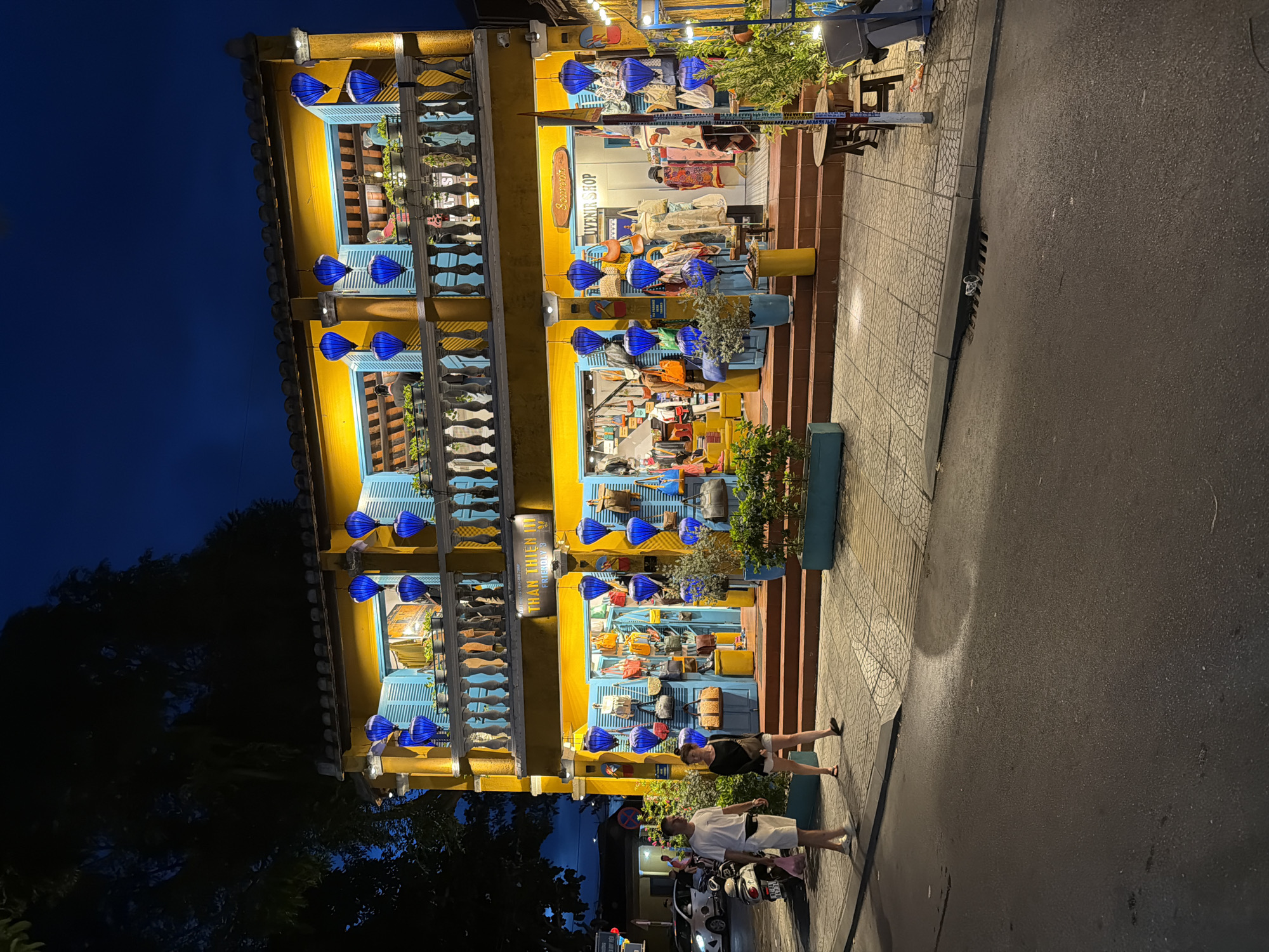 In the evening, a two-story yellow building with blue shutters and balconies is illuminated. Blue lanterns hang from the balconies and in front of the shop on the ground floor. The shop appears to be a souvenir shop, with bags and clothing displayed in the windows and outside. Two people, a man and a woman, are walking on the sidewalk in front of the shop. A white scooter is parked to the left of the shop. The street in front of the building is wet, suggesting it may have recently rained. The scene is set in the historic city of Hoi An, Vietnam, a UNESCO World Heritage site known for its well-preserved ancient town. The overall ambiance is lively and atmospheric, characteristic of Hoi An's vibrant street life after dark.
