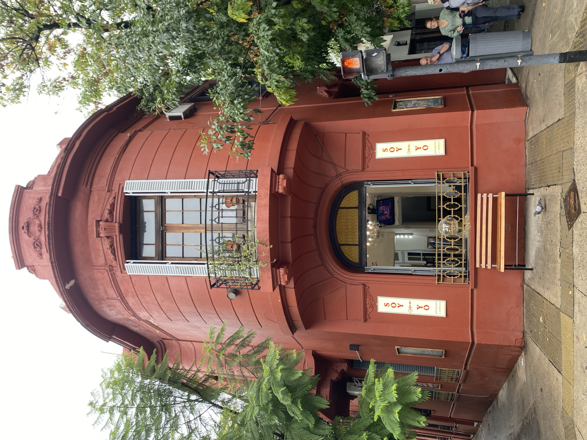 The image captures the facade of a reddish-brown, ornate building located in Buenos Aires, Argentina. The building has a distinct curved architectural design with a window featuring white shutters and a balcony. A large arched window is positioned above a doorway with an elaborate wrought iron gate. Signs reading "SOY YO Peluqueria de Artistas" are visible above and below the window. A traffic light is attached to the building's facade. There are a couple of people next to a garbage can on the street. Green foliage partially obscures the view of the building. The sidewalk is adjacent to the building, and the perspective is slightly angled upwards.