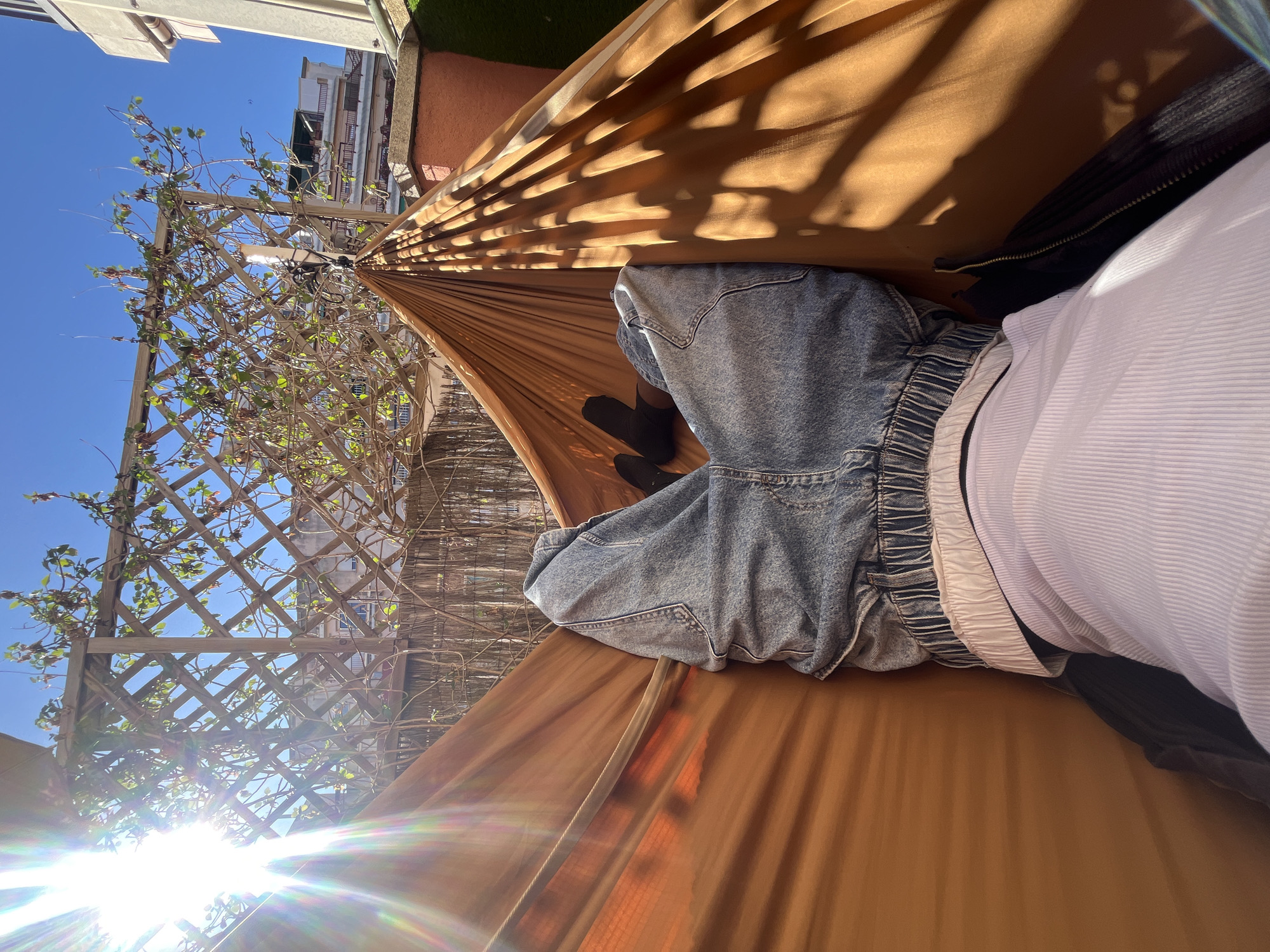 A person is lying in a hammock under a patio awning, wearing blue jeans, black socks, a dark jacket, and a striped white shirt. The hammock is suspended near a lattice fence with climbing vines. The location appears to be in Barcelona, Spain, based on the architecture and vegetation visible in the background. The sky is clear and sunny, with sun flares visible at the bottom left of the frame.