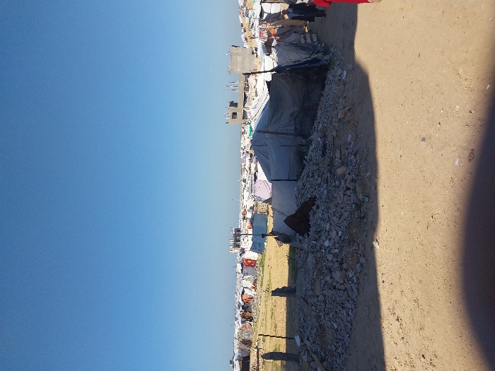 The image depicts a wide-angle view of a densely populated, improvised settlement under a clear blue sky in Khan Yunis, Palestinian Territory. In the foreground, a sandy dirt path is partially shaded by an unseen object to the left. To the left of the path, a substantial pile of concrete rubble and stones sits, with three broken concrete posts vertically protruding behind it. The midground features numerous makeshift shelters constructed from tarpaulins, fabric sheets, and various repurposed materials, supported by wooden poles, extending towards the background. A partially visible individual in a red top and black pants walks away from the camera along the path on the right, followed by another person in dark clothing. In the background, an extensive vista of similar temporary shelters stretches across the horizon, interspersed with several incomplete, multi-story concrete structures in various stages of construction or disrepair. The sky above is uniformly clear and bright blue.