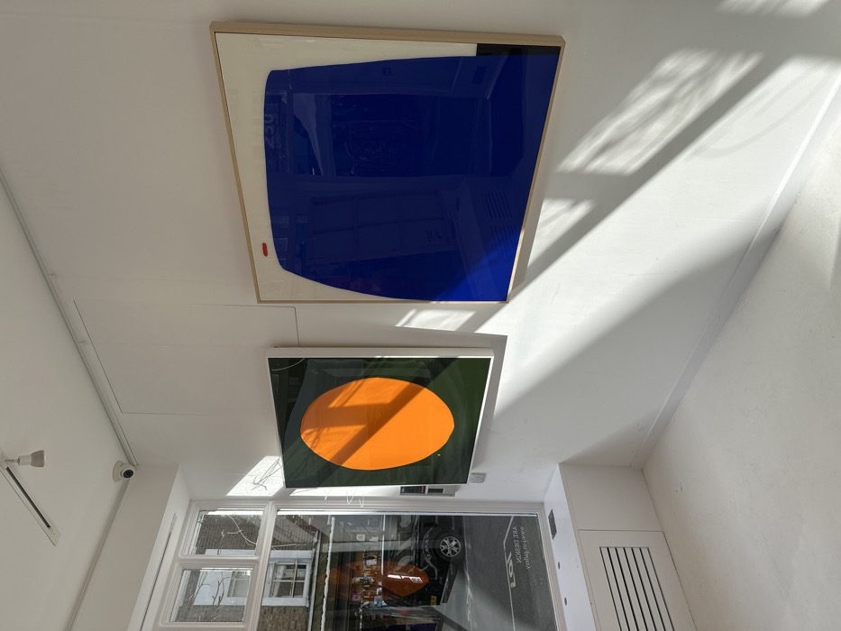 The image depicts the interior of M-U Gallery, located at 107 Portobello Road, London W11 2QB, United Kingdom. The space features white walls, a white floor, and a white ceiling equipped with track lighting and a white CCTV camera. On the left, a multi-pane window offers a view of the street, reflecting a black vehicle, brick buildings, and a white canopy. Reflected signage, "ART DESIGN" and "www.m-u.gallery", is visible on the lower window pane. A white radiator cover with a horizontal grille is positioned beneath the window.
Two abstract artworks are displayed on the white wall. The left artwork is a vertically oriented rectangular piece featuring a large orange oval on a dark green background, framed in white. The right artwork, a square piece in a light wooden frame, presents a large, glossy royal blue, irregularly curvilinear form against an off-white background. This blue form is accented by a small red horizontal dash near its top left and a small black vertical rectangle at its bottom right. Reflections of the exterior scene are visible on the blue surface. Sunlight streams through the window, casting distinct geometric shadows across the interior walls and floor. No people are visible within the scene.