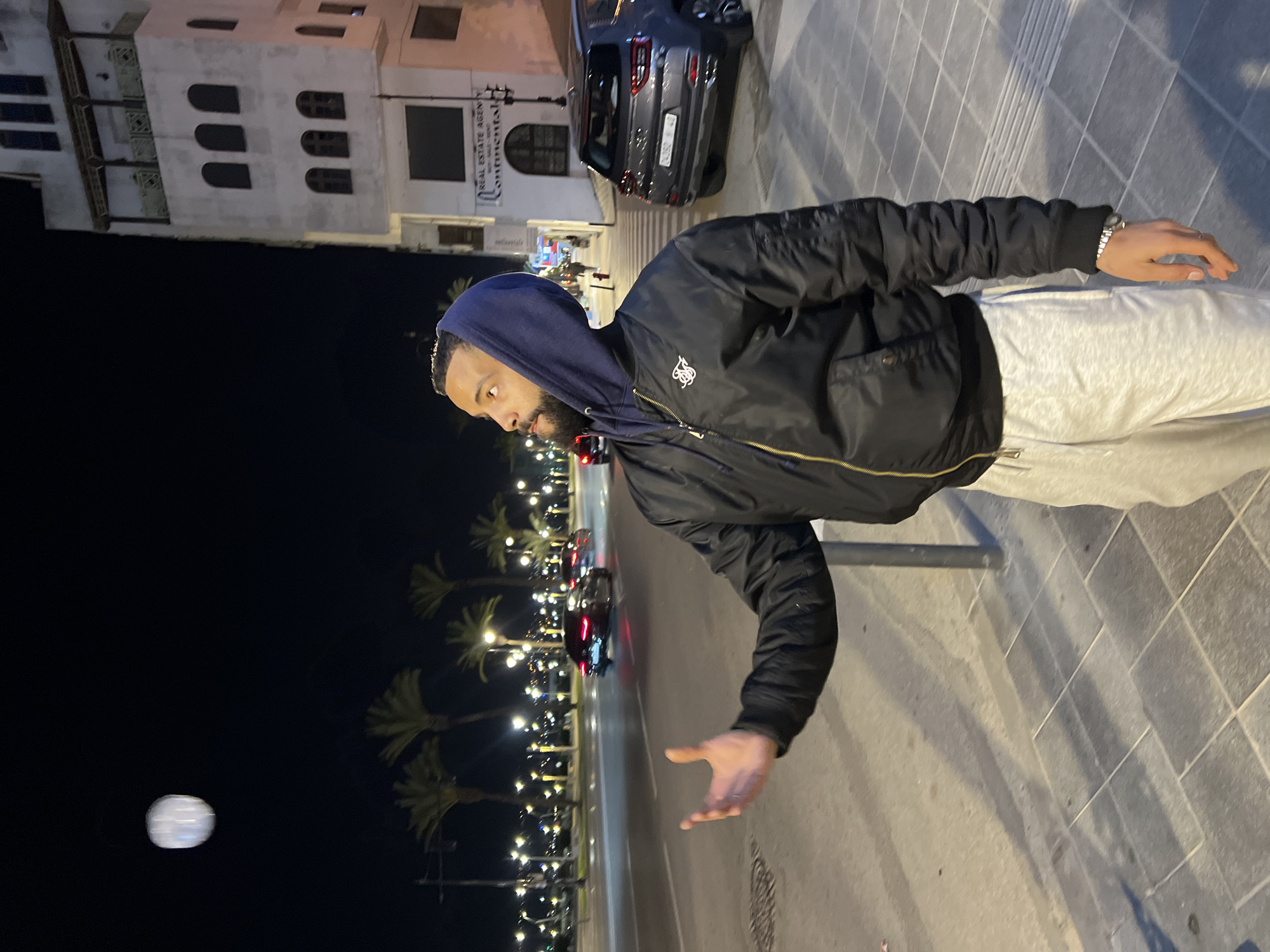A nighttime scene depicts a male individual on a paved sidewalk alongside a multi-lane road in Tanger, Morocco. The man, with a beard and short dark hair, is positioned in the mid-ground, facing left and looking upwards. He wears a navy blue hooded sweatshirt under a black bomber jacket with a gold zipper and a "SIKSILK" logo on the left chest, paired with light grey sweatpants. His right arm is raised with an open palm, directed towards a blurry, luminous white, ovoid object suspended in the air above him. His left arm is down by his side, with a silver watch visible on his wrist.
Behind the man, a wide asphalt road extends with multiple lanes. Several vehicles with illuminated taillights are visible, suggesting ongoing traffic. The road is lined with palm trees and numerous streetlights, which cast light onto the scene and create reflections on the wet-appearing road surface. In the background, across the street, a parked dark grey SUV with a Moroccan license plate (D3350F 48) is visible. Further back, a multi-story white building with arched windows and balconies features signs on its ground floor reading "REAL ESTATE AGENCY" and "Continental." Distant city lights are visible across the horizon, possibly indicating a coastal view. The sidewalk consists of grey square and rectangular pavers.