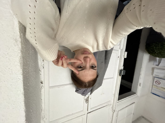 This image captures a young female indoors in Strasbourg, France. She is depicted from the chest up, looking directly at the camera with a neutral expression, and exhibits a noticeable reddish-brown discoloration or bruise beneath her right eye. Her brown hair is pulled back from her face. She wears a cream-colored, high-necked knitted sweater featuring small perforated patterns on the sleeves. Her left hand (viewer's right) is raised, displaying a 'peace' sign with her index and middle fingers, and her fingernails are adorned with red and white glitter. The background consists of white walls and built-in, paneled white cabinetry above an open doorway, which leads into a dark, unlit area. To the far left, a portion of a green topiary-style plant in a dark pot is visible, along with several documents pinned to the wall, one displaying a blue border and a red 'no' symbol. A grey jacket hangs on a hook to the right of the doorway.