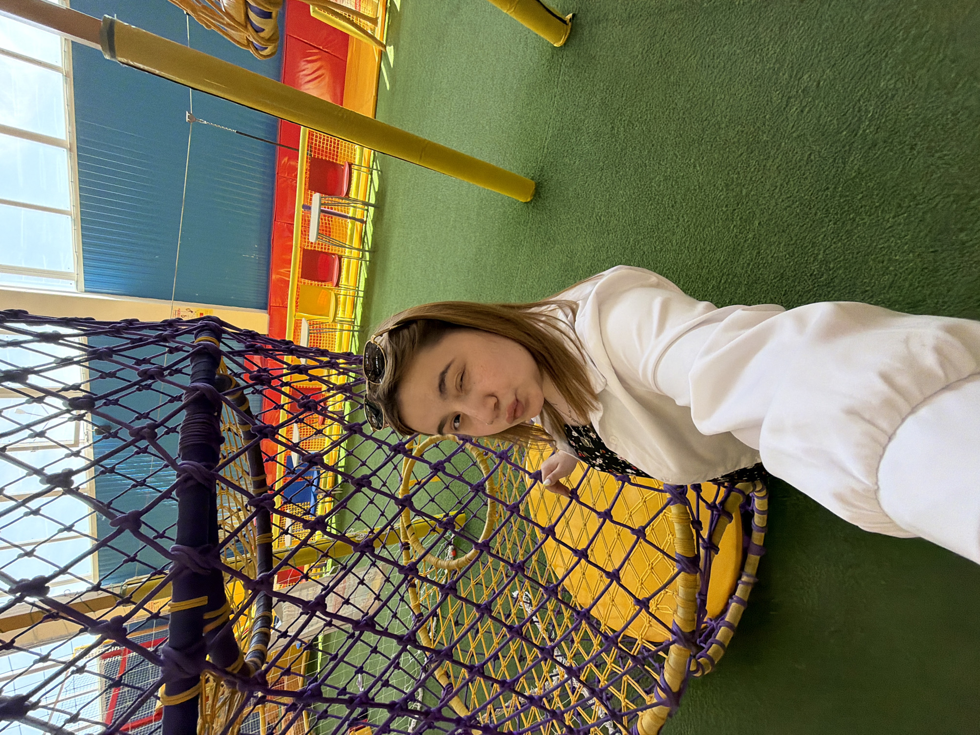 A person in a white jacket is standing next to a purple and yellow rope structure. The location appears to be an indoor play area, possibly in Chaikovsky, Russia, given the prompt. The background includes green flooring and brightly colored play equipment.
