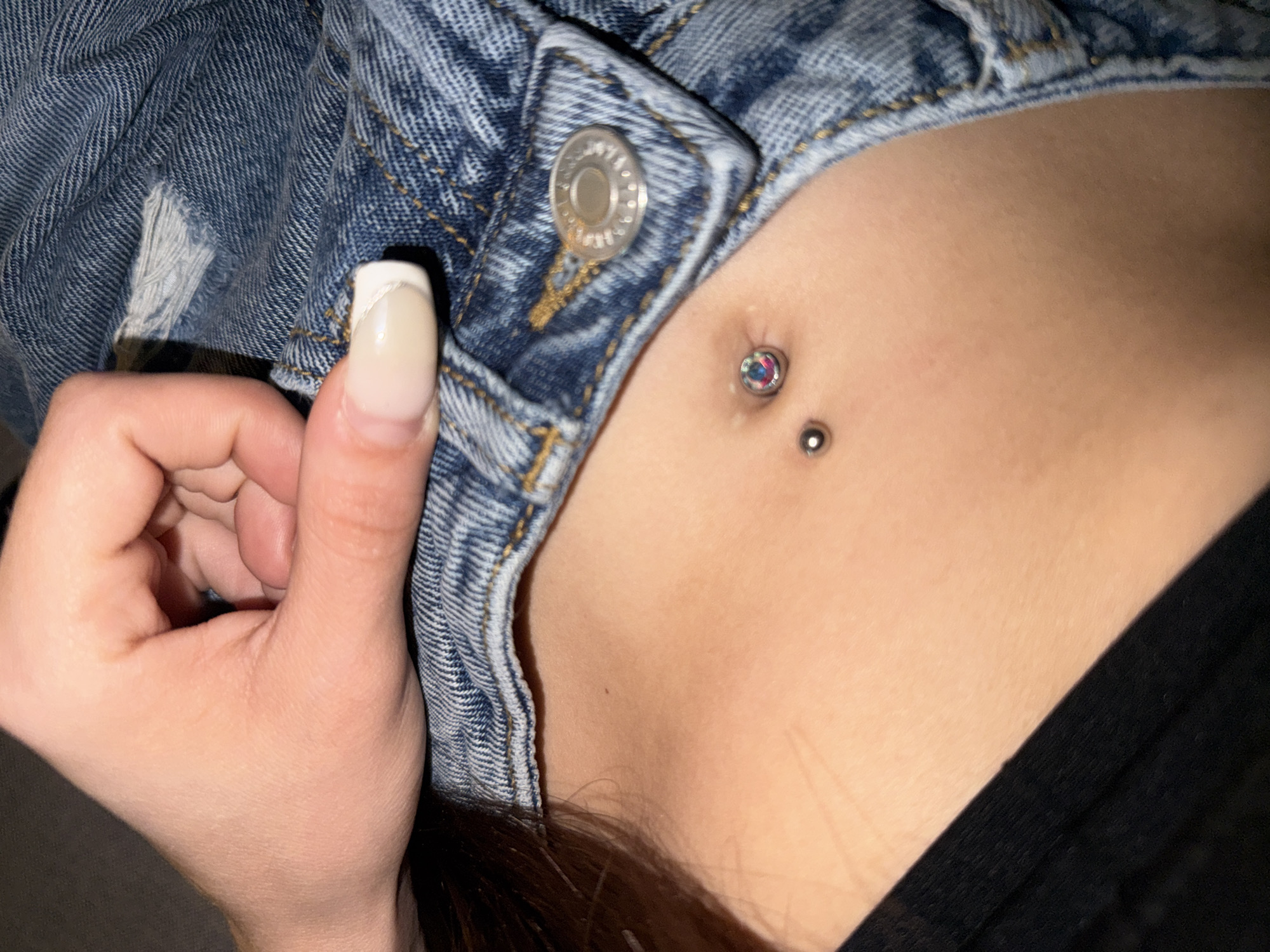 A close-up, overhead shot captures a person's torso, specifically their midriff, adorned with a navel piercing. The piercing features a top ball and a decorative bottom gem with iridescent colors. Above the piercing, a portion of denim clothing, possibly a jacket, is visible with a prominent button. A hand with neatly manicured nails rests on the denim, obscuring part of the upper torso and head. The background is dark and out of focus. The overall impression is intimate and personal, with the focus drawn to the piercing and the texture of the denim.