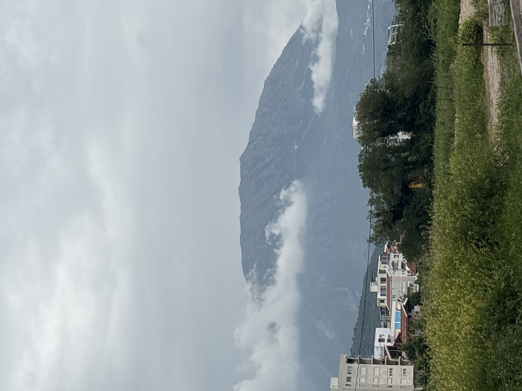 The photograph depicts a mountainous landscape scene in Bar, Montenegro, under an overcast sky. The dominant feature is a large mountain, its peak obscured by low-hanging clouds. The mountain's rocky texture is visible despite the cloud cover. In the foreground, there are several white buildings with red and blue rooftops. These buildings appear to be residential. Adjacent to the buildings is a patch of dense green vegetation. The overall impression is one of a serene but cloudy day in a mountainous region, likely on the outskirts of Bar.