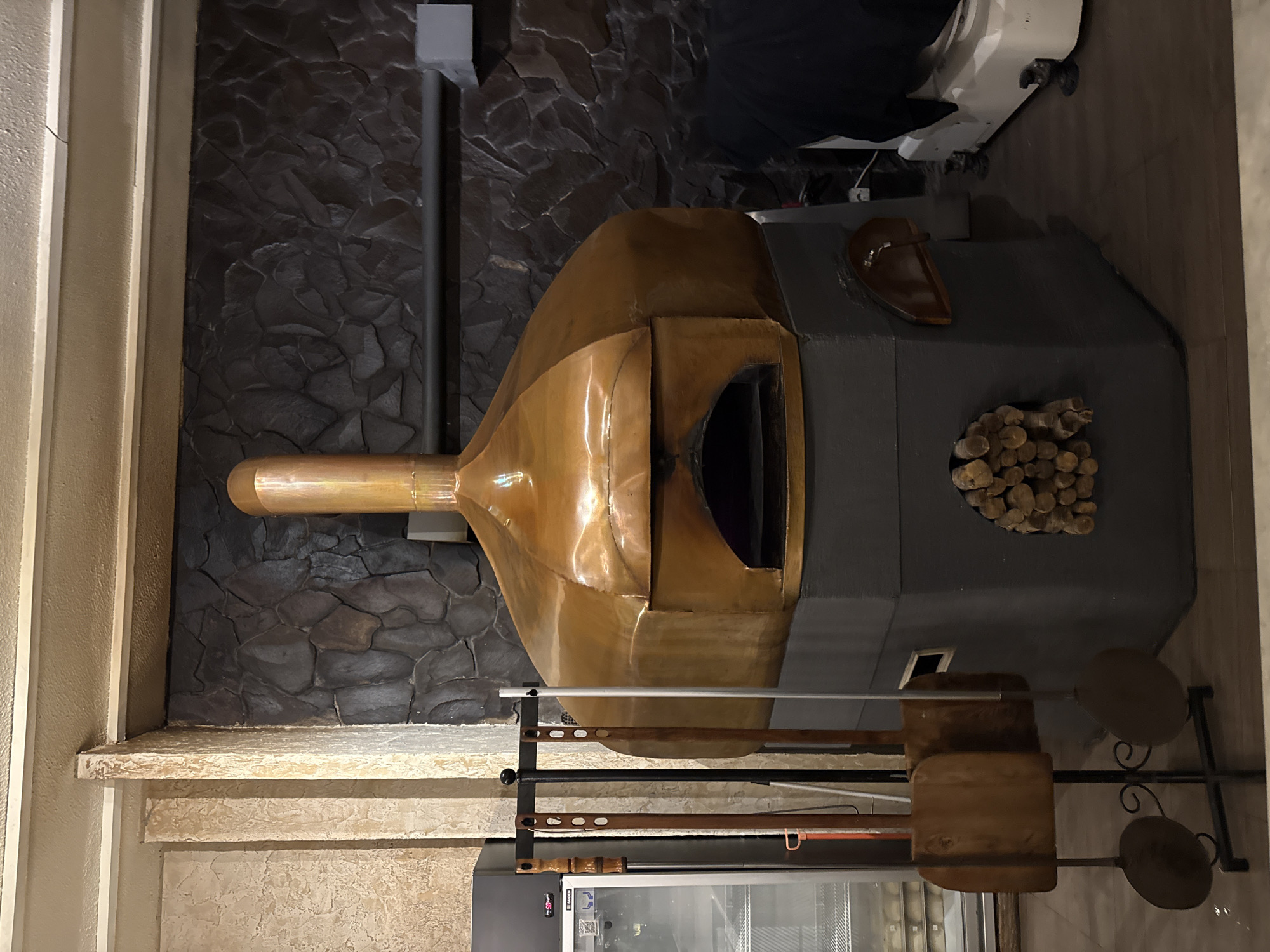 The image displays an elaborate indoor setup, likely in a restaurant or commercial kitchen, centered around a large, distinctive pizza oven. The oven features a prominent, polished copper dome and a matching chimney pipe extending upwards. This copper structure is integrated into a robust dark gray base, possibly made of stone or concrete, which houses the oven's wide, dark opening.
To the right side of the gray base, a circular recess holds a neatly stacked pile of small, cut firewood logs. Further right on the base, a dark, handled pan or tray rests. The oven is positioned against a dark, textured wall, resembling rough-hewn stone or slate. To the left of the oven, a lighter-colored wall or architectural column is visible. Above and slightly behind the oven, some white kitchen equipment, possibly a mixer or another large appliance, is partially visible.
Below the oven, a metal stand is equipped with various long-handled tools, including several pizza peels, some with wooden blades and handles, and others appearing metallic. Further to the right, a decorative metal stand holds two dark, circular elements, which could be small serving tables or decorative trays. To the far left, at the bottom, a portion of a commercial refrigerator or cooler is visible, with the brand name "BOSCH" faintly legible on a small red label. No people or specific activities are depicted. The lighting is artificial, and the scene presents a blend of rustic and refined elements, characteristic of a professional culinary environment.