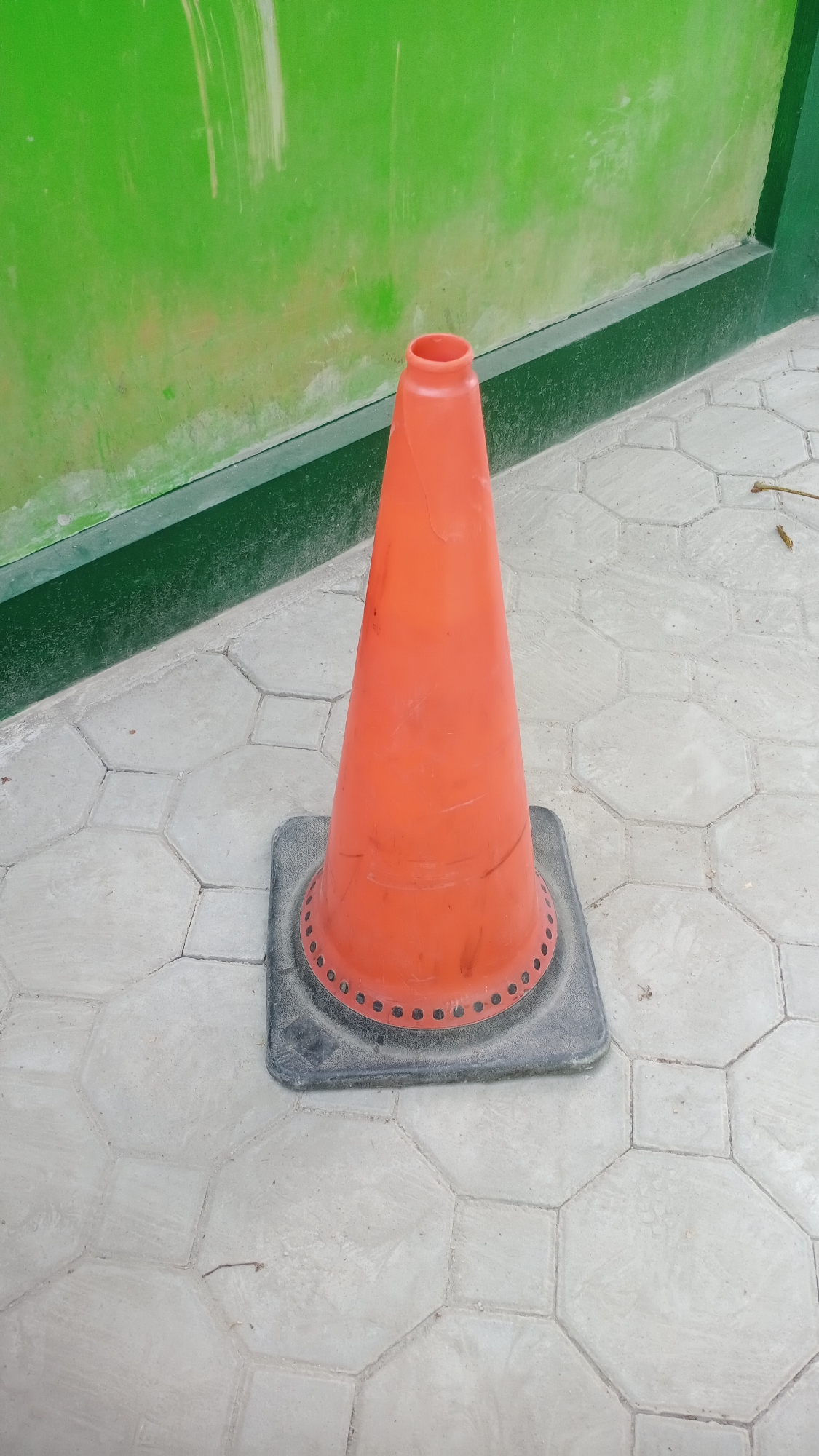 A single orange traffic cone with a black base stands on a patterned tiled surface. Behind the cone, a green wall with some vertical white marks is visible, bordered by a dark green band. The scene is located outdoors on a tiled walkway, possibly in Joho, Indonesia, given the context provided. There are no people or other objects prominently featured in the image.