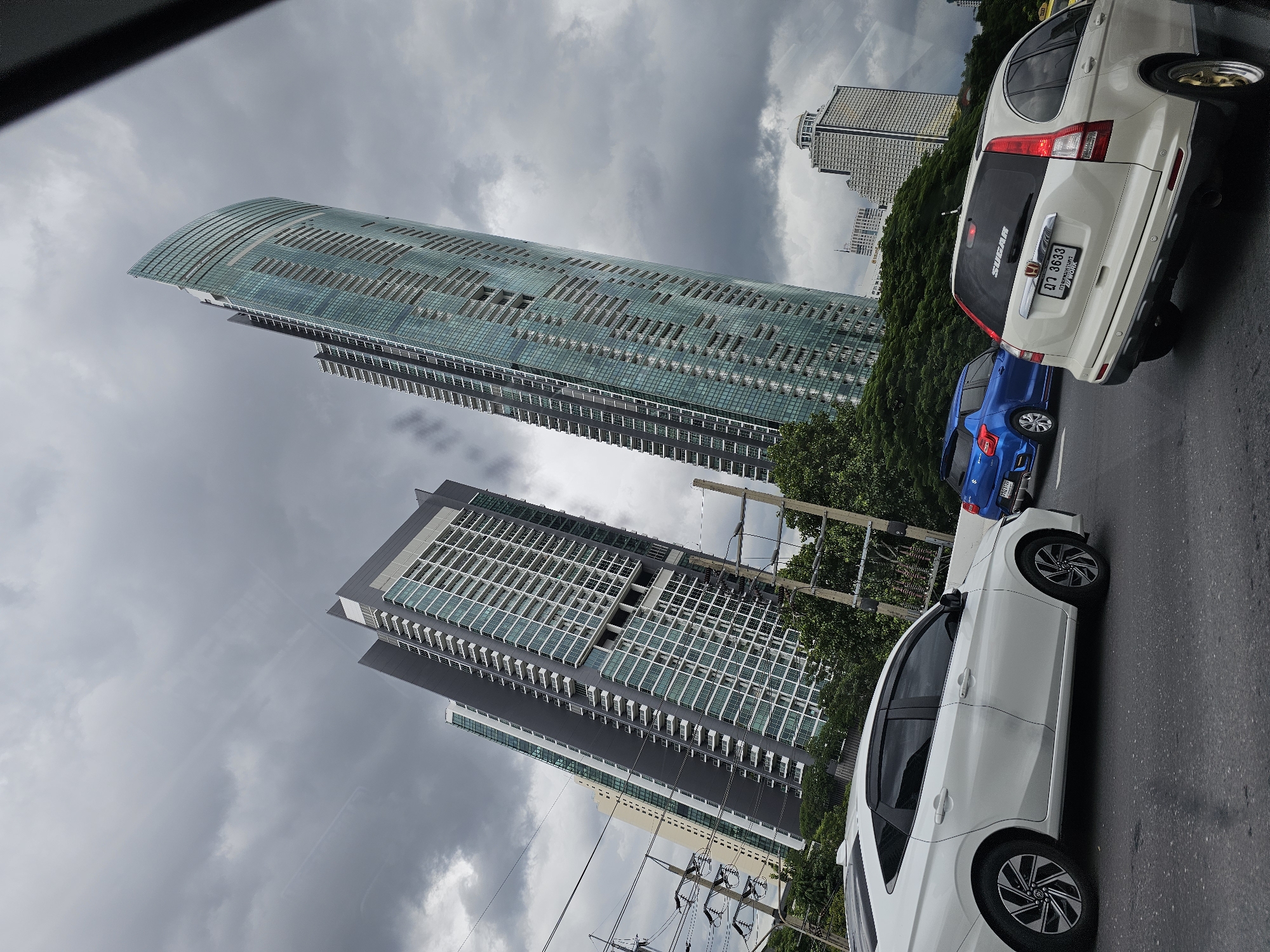 The image captures a street-level view of a cloudy day in Bangkok, Thailand, featuring modern architecture and traffic. In the foreground, a white sedan is on the right, and a blue hatchback is to its left. A white SUV is positioned in the right lane, its rear facing the camera. In the background, two prominent skyscrapers dominate the skyline: one with a distinctive curved, glass facade in teal hues, and another adjacent, taller building with a gray exterior and many windows. Further in the distance, other buildings and structures are visible, including what appears to be a hotel with a domed roof. Electrical wires and poles line the side of the road, and lush green trees separate the road from the buildings. The sky is overcast with dark, heavy clouds, suggesting an impending storm. The overall impression is one of an urban environment in Thailand, characterized by contemporary high-rise buildings and the presence of vehicular traffic on a cloudy day.