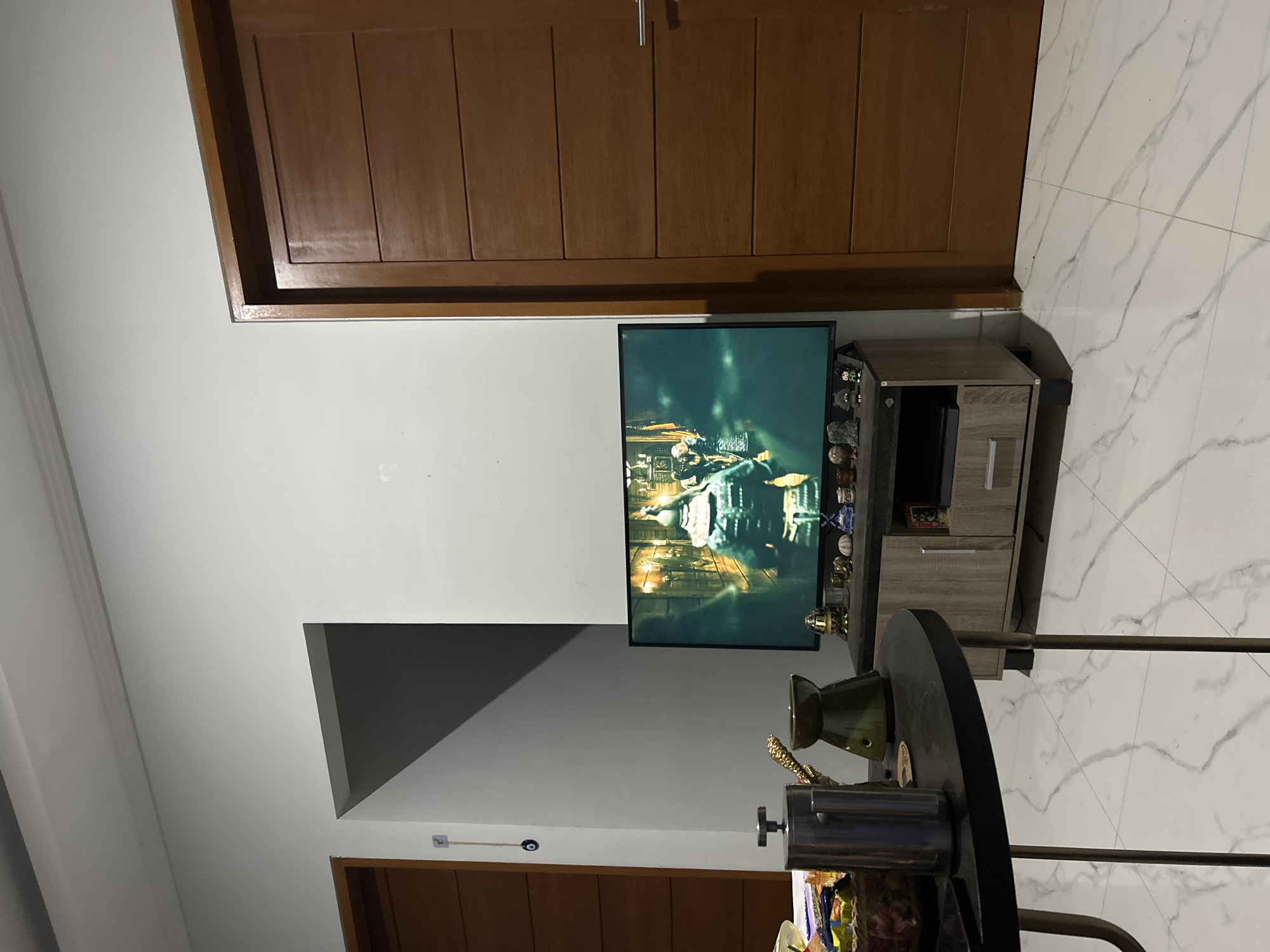 A well-lit, indoor scene shows a living room area. On the left, a wooden door is partially visible. A dark, round table with metal legs stands in the foreground. On the table, a French press coffee maker and a green ceramic diffuser are prominent. The background features a large flat-screen television displaying a scene from a historical drama, likely a movie or series. The television is placed on a light brown media console. To the right, a closed wooden door with horizontal panels dominates the right side of the frame. The floor is tiled with a white and grey marble pattern. The wall is painted white. The overall impression is one of a comfortable and modern living space. The image is taken in Sukawati, Indonesia.