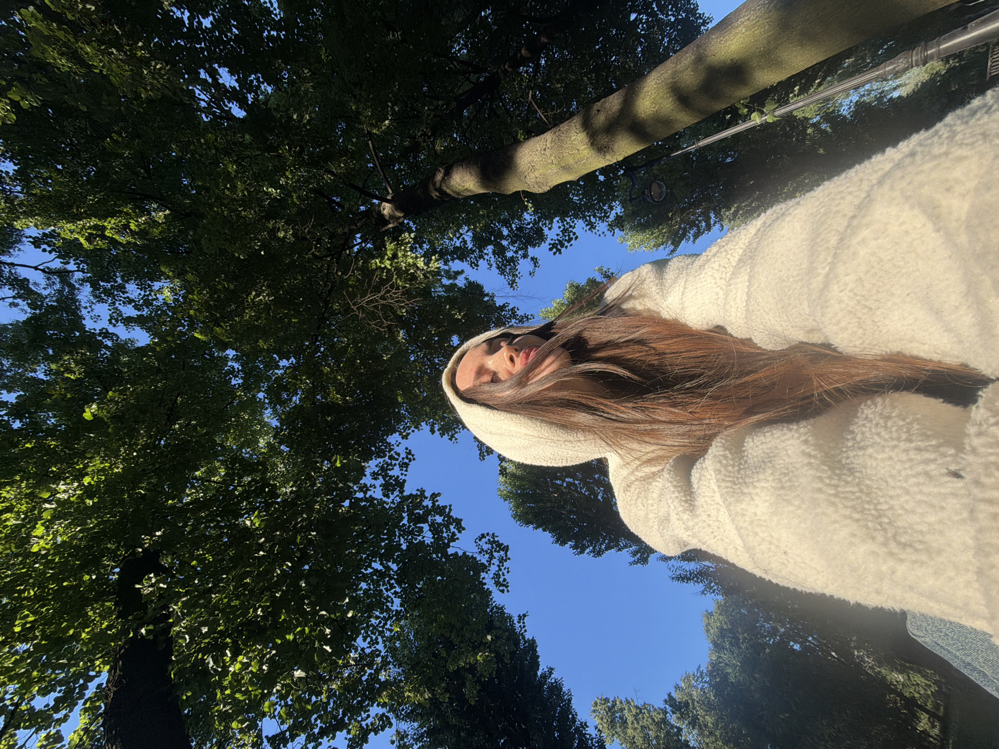 A low-angle, full-body shot captured from behind shows a young woman with long, dark brown hair wearing a cream-colored fleece hooded jacket and a denim jacket underneath, looking upwards towards the sky. She is positioned in the foreground, with her face partially obscured by her hair and hood, and her arms are bent and out of view. The background consists of a clear blue sky and tall green trees. The lighting suggests it is daytime, with sunlight filtering through the leaves, creating dappled shadows on the tree trunks and the woman's clothing. The image is likely taken in an outdoor urban park setting. Based on the visual elements, it is not possible to definitively determine the specific location within Saint Petersburg, Russia.