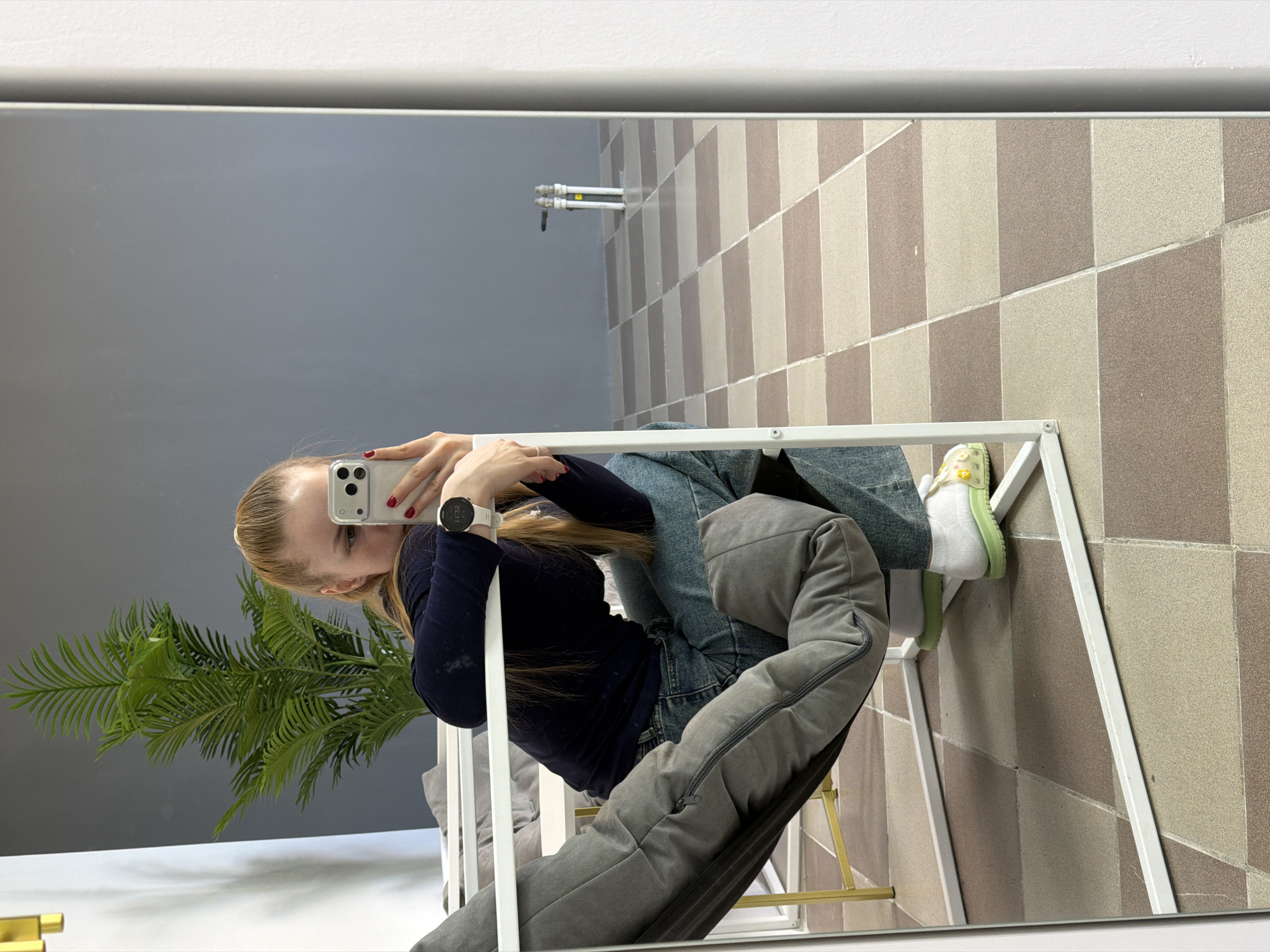A reflection of a person taking a selfie in a mirror, presumably in Berezniki, Russia. The person is seated, wearing jeans, a dark long-sleeved shirt, white socks, and green and yellow footwear. The reflection includes a glimpse of a plant, a gray cushion, and a tiled floor. Pipes are visible in the background. The person is holding a white smartphone with visible cameras. A smartwatch is on the wrist of the arm holding the phone. The mirror has a white frame. The scene appears to be indoors.