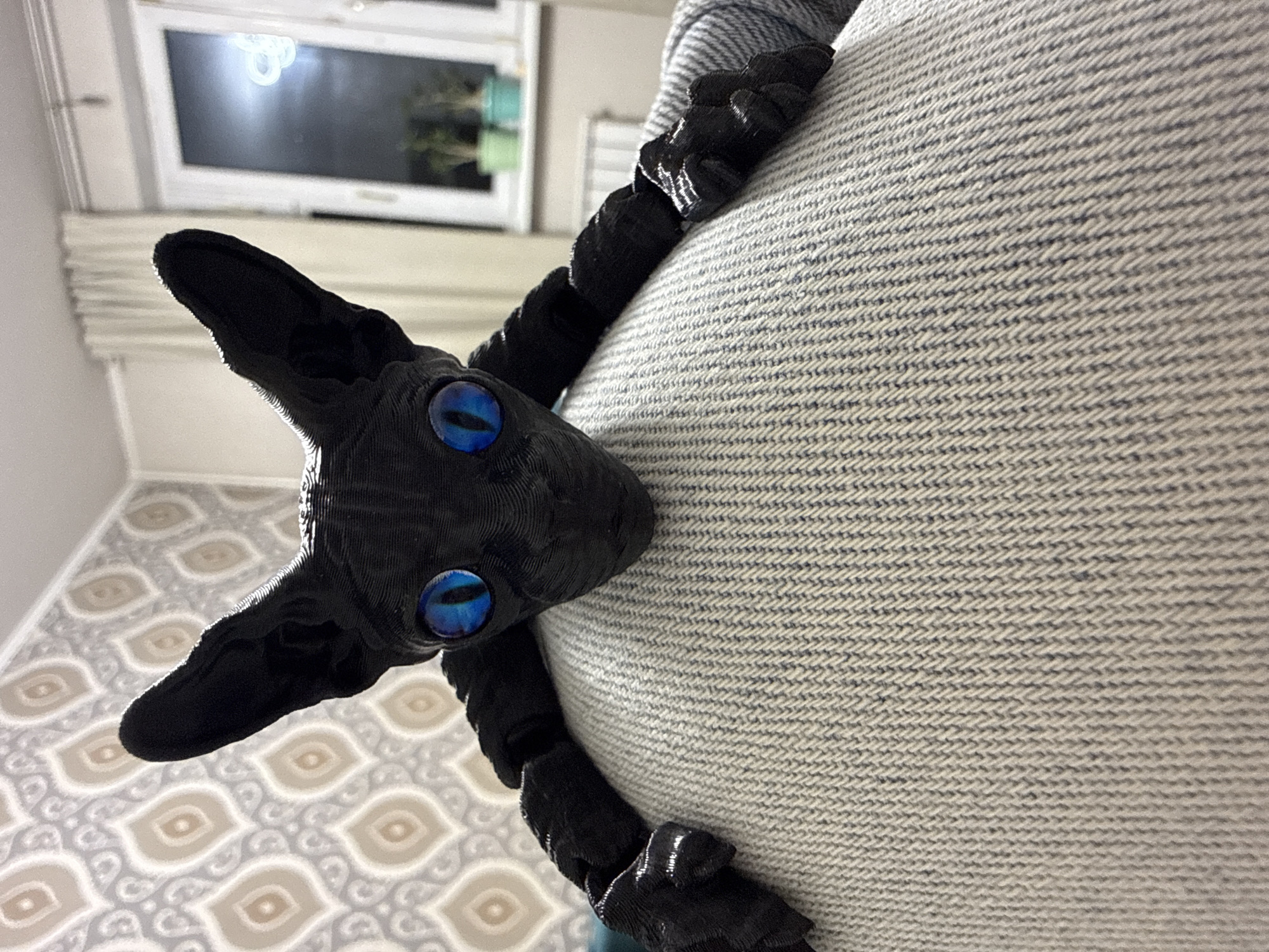 A black, articulated, cat-like figure with blue eyes is positioned against a light gray, horizontally striped surface. The figure has pointed ears and is segmented, suggesting it may be a toy or model. The surface it leans against appears to be a textured fabric.
In the background, a window with a white frame is visible. The interior setting suggests a domestic environment, with a patterned floor in the lower left corner. There are no people visible.
There is no indication of this image being taken in Khimki, Russia.