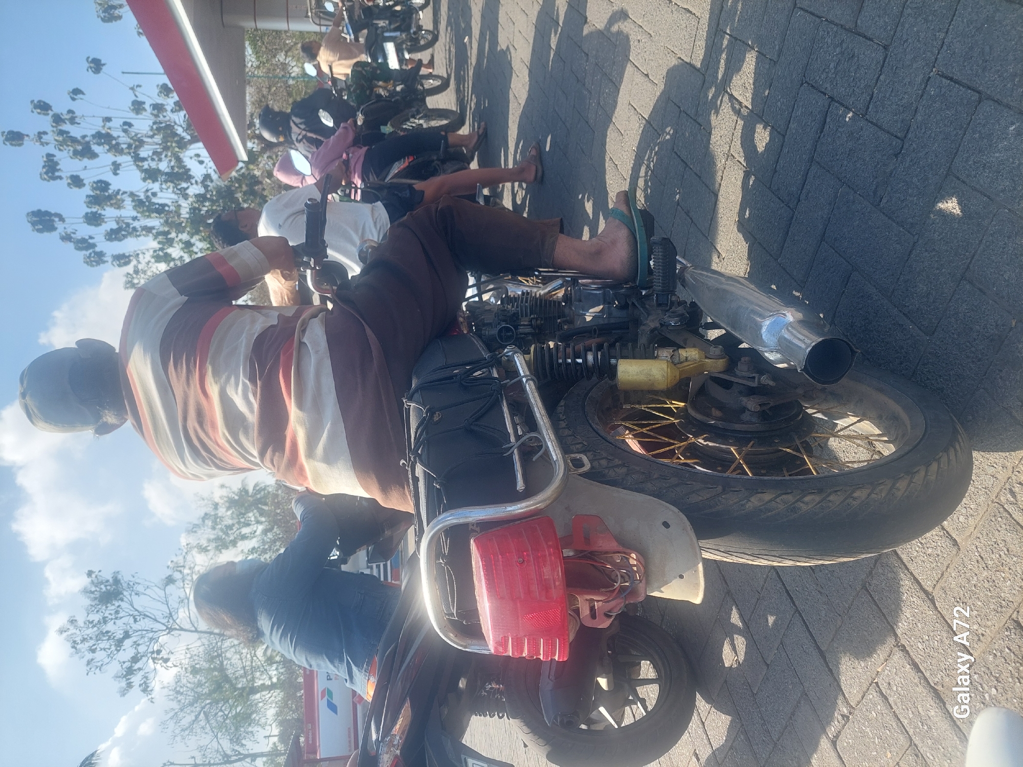 This image depicts a scene at a gas station in Sengguruh, Indonesia, with several motorcycles lined up. In the foreground, a man wearing a striped shirt and brown pants is seated on a motorcycle, with the rear of the motorcycle prominently displayed. The motorcycle features a red taillight and a chrome exhaust pipe. Behind him, other individuals are on motorcycles, some wearing helmets. A woman in a denim jacket is visible on a motorcycle to the left. The gas station canopy, with a red trim, is visible in the background. The ground is paved with interlocking brick-like stones, and the sky is blue with some clouds. The presence of multiple motorcycles suggests a queue or a gathering at the gas station.