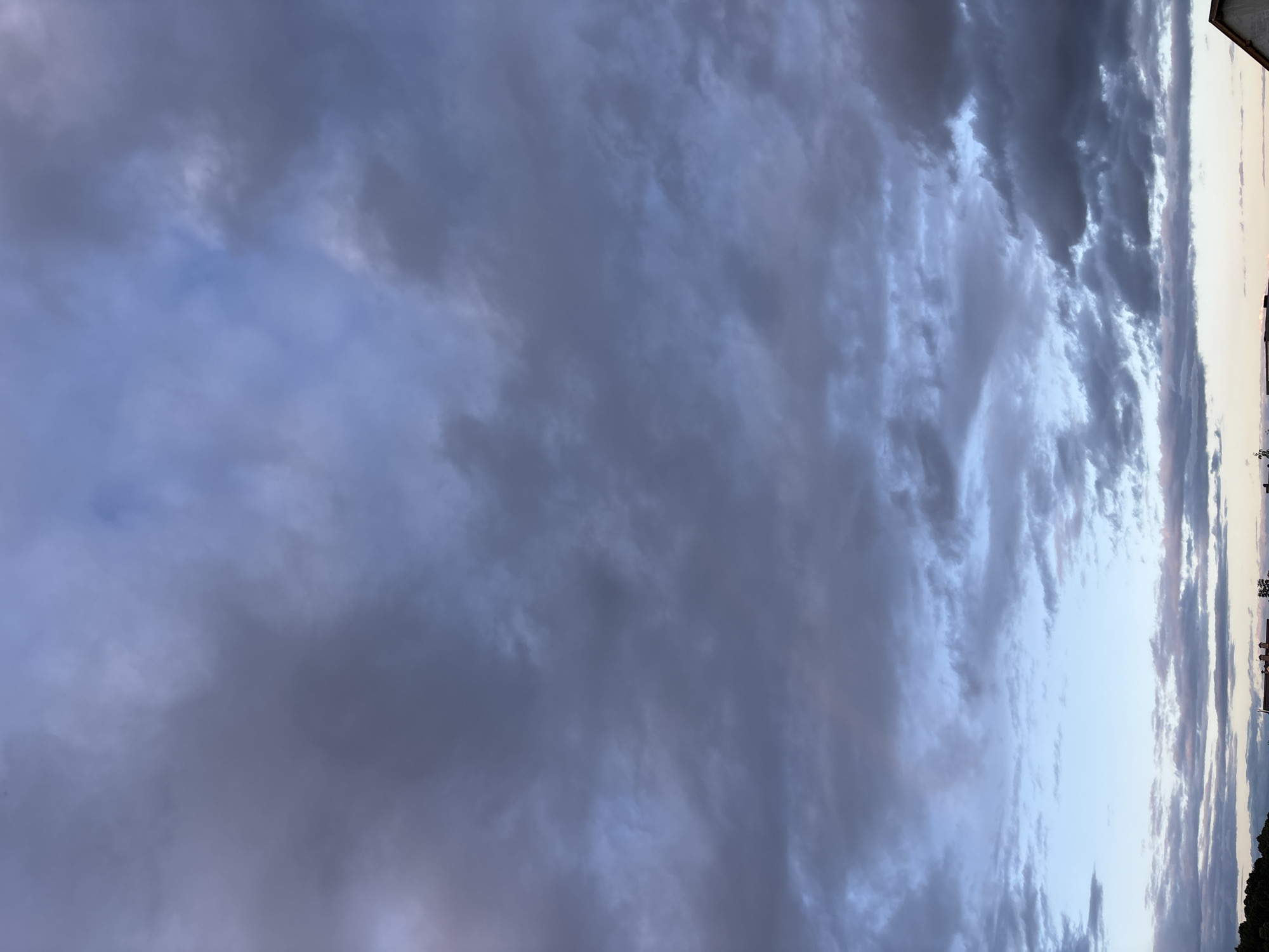 The image depicts a cloudy sky over Zvenigorod, Russia. Dark, ominous clouds dominate the upper portion of the frame, with streaks of lighter blue and hints of pink suggesting either a pre-dawn or post-sunset light. Below the dense cloud cover, a clearer patch of pale blue sky is visible towards the horizon, interspersed with thinner, wispy clouds. The lower edge of the image shows a silhouetted treeline on the far left, followed by the rooftops of buildings. One building clearly displays a red roof with chimneys. The overall impression is of a moody, overcast sky, potentially indicating approaching rain or the aftermath of a storm. No people or specific actions are discernible in the image.