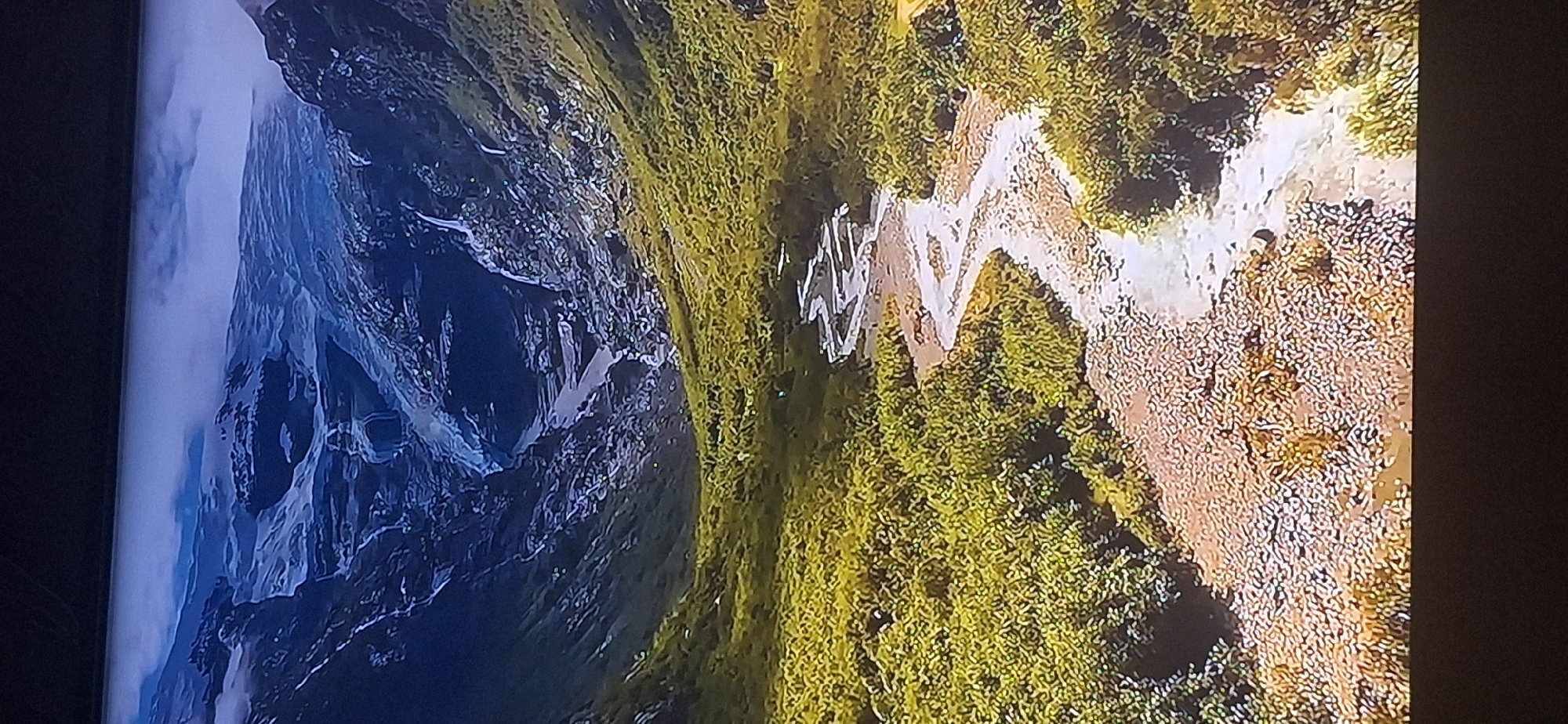 The image depicts a vast, mountainous landscape with a winding river flowing through a valley. The upper portion of the image shows a glacier or snow-covered mountain range with cascading waterfalls. As the landscape descends, it transitions into steep, rocky slopes covered in green vegetation. The river, appearing murky and braided, cuts through the valley floor, which is densely vegetated with trees and shrubs. The overall scene conveys a sense of rugged natural beauty and isolation. There is no indication of the city of Bila Tserkva, Ukraine, in this image.