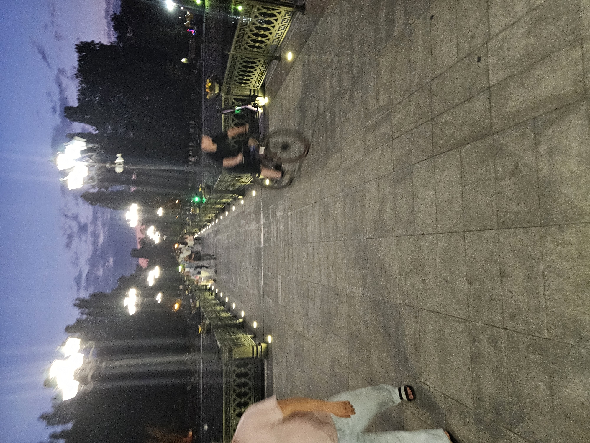 The image depicts a nighttime scene on a bridge or elevated walkway in Cherkessk, Russia. The walkway is paved with large, square tiles and lined with ornate metal railings. Numerous streetlights, in a vintage style, illuminate the pathway. In the background, silhouetted trees form a dark line against a twilight sky with scattered clouds.
Several people are present on the walkway. On the left side, a person wearing a light pink t-shirt and light-colored pants, with sandals, is partially visible and out of focus, suggesting movement. Further along the walkway, a group of people are walking in the distance, also appearing somewhat blurred due to motion or low light conditions. In the middle right of the image, a person is riding a bicycle, their form heavily blurred by motion. Beside the cyclist, an electric scooter is parked. The overall impression is of a public space being utilized by pedestrians and a cyclist during the evening.