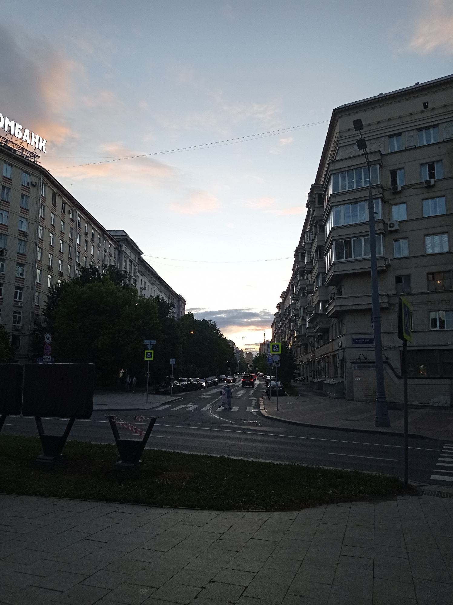 The image depicts a street scene in Moscow, Russia, at dusk. The foreground shows a grey paved sidewalk with a strip of grass bordering a street. On the left side of the street, there is a large, multi-story residential building with many windows. A large neon sign that reads "ОМБАНК" is visible at the top of this building. Across the street, another large residential building of similar architectural style lines the right side of the road. Several cars are parked along the curb on the right, and traffic is visible on the road. Pedestrians are crossing the street at a crosswalk. The sky is partly cloudy with hues of pink and orange from the setting sun. A street lamp stands near the right-side building.
