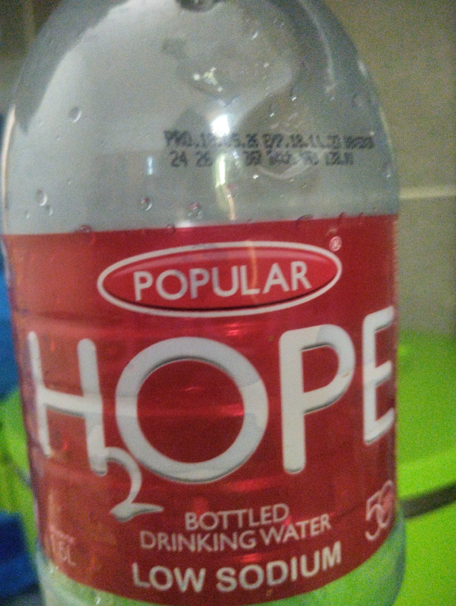 This media file is a close-up, eye-level shot of a plastic water bottle with a red label. The bottle appears to be partially filled with water, as evidenced by condensation droplets on its surface. The label is predominantly red and features white text.
The most prominent text on the label reads "POPULAR" within an oval shape, and below that, the word "HOPE" in large, bold, white lettering. Further text indicates "H₂O", "BOTTLED DRINKING WATER", and "LOW SODIUM". There is also a smaller number "50" visible on the right side of the label. In the upper section of the bottle, above the red label, blurry black text is visible, possibly a date stamp or manufacturing code. This text appears to read "PRO.128.05.25 EXP.18.11.23 18.25 24 26".
The background is out of focus, but appears to be a bright green, textured surface. The lighting suggests it is indoors, possibly in a kitchen or pantry setting. No people are visible in the image, and no specific activity is taking place other than the bottle being present. The overall impression is of a product shot, highlighting the branding of the bottled water.