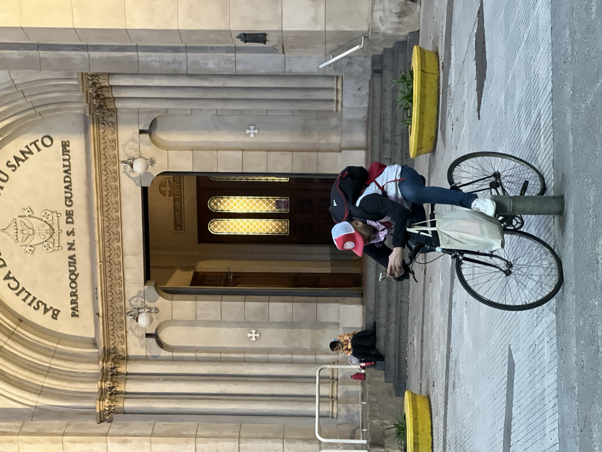 The image shows the Basilica of the Holy Sacrament in Buenos Aires, Argentina. The basilica's façade is predominantly stone with arched entrances and detailed carvings. On the right, two individuals are depicted; one is leaning over, possibly interacting with a bicycle that is propped up against a pole. Another person is sitting on the steps near the entrance, seemingly observing or resting. Yellow plant holders are visible near the steps.