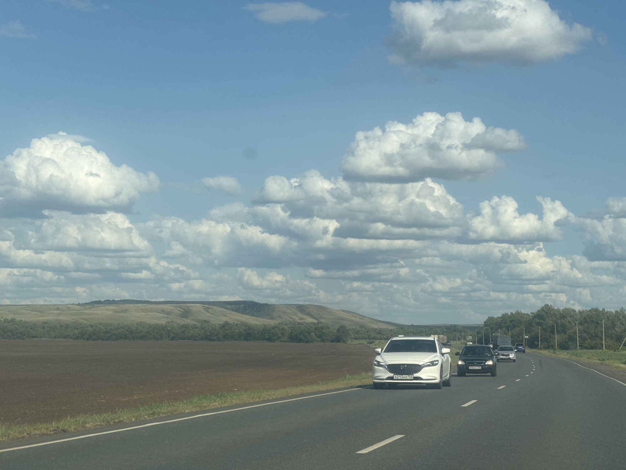 The image depicts a highway scene on a sunny day with a blue sky filled with fluffy white clouds. In the foreground, a white car with license plate A213AB 156 is driving on the right side of the road. Several other vehicles are visible further down the highway, including a black car and a truck. The landscape beside the highway features a cultivated field, a line of trees, and rolling hills in the distance. The road appears to be a rural highway, consistent with a location like Novosergiyevka, Russia. There are no people visible in the image.