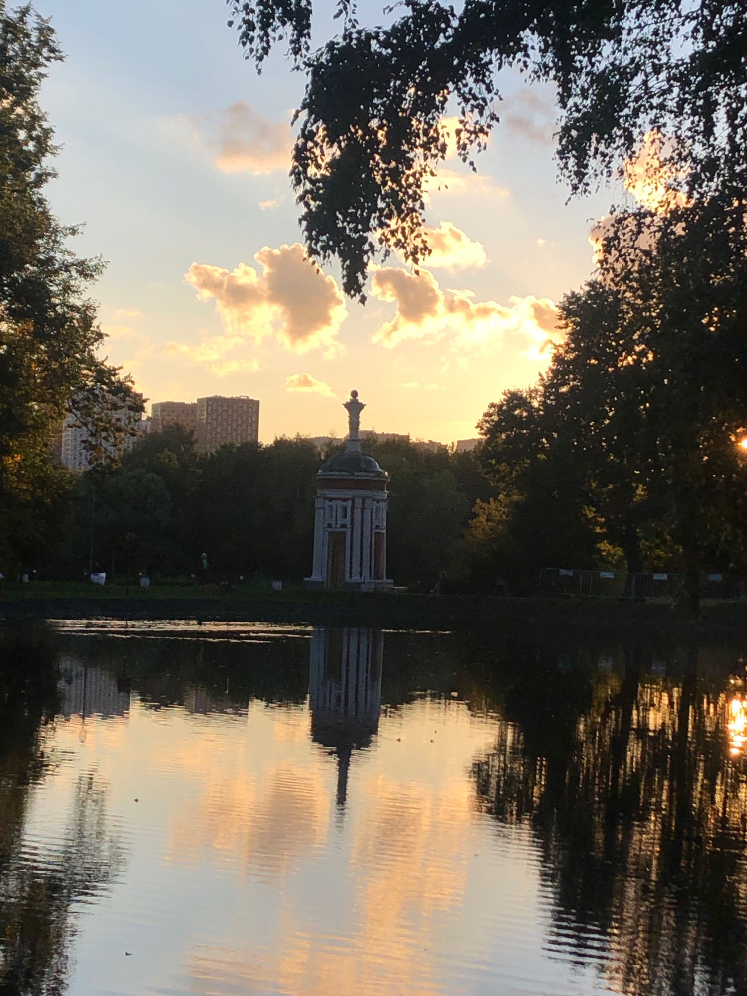 The image depicts a tranquil scene at sunset in the city of Khimki, Russia, likely at the Arkangelskoye Estate. A still body of water, reflecting the warm hues of the setting sun and the sky, dominates the foreground. The water's surface is gently rippled, with the reflections of trees, clouds, and a prominent architectural structure.
In the mid-ground, a white, classical-style pavilion with red accents stands near the water's edge, its symmetrical form mirrored in the water. Behind the pavilion, a dense line of trees creates a dark green backdrop, transitioning into distant apartment buildings visible on the horizon. The sky above is a blend of soft blue, illuminated by the golden and orange light of the setting sun, with scattered clouds catching the light. Silhouettes of trees frame the top of the image, their leaves and branches partially obscuring the sky. A few small, dark shapes, possibly birds or ducks, are visible on the water. The overall atmosphere is one of serenity and natural beauty at dusk.