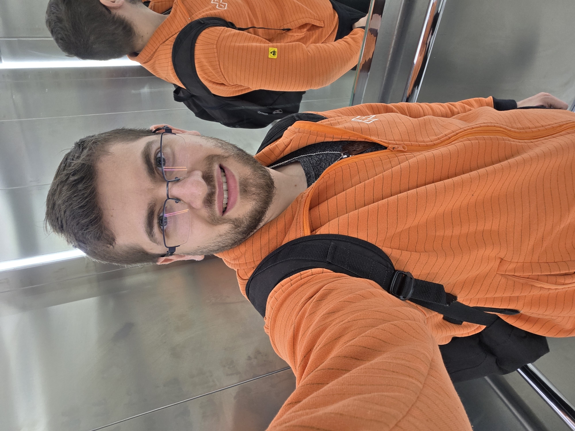 A man with brown hair and a beard wearing glasses and an orange zip-up hoodie with a black backpack is taking a selfie in an elevator with a mirrored wall. The location is presumed to be Plovdiv, Bulgaria, based on the prompt's instructions. The elevator walls are stainless steel. He is smiling slightly. His reflection is visible in the mirror. The lighting appears to be fluorescent.