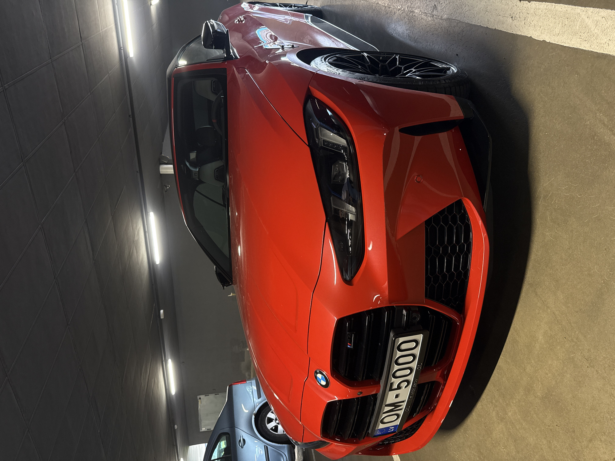 The image shows a red BMW car parked in an indoor parking garage. The car has a license plate that reads "OM-5000" with "LV" printed, indicating that the vehicle is registered in Latvia. The presence of the Latvian license plate suggests that the photo was taken in Latvia. However, there's no specific information to confirm if this image was taken in the city of Riga. The garage itself is lit by fluorescent lights. Other cars can be seen parked in the background. The image appears to have been taken at a close range, focusing on the front of the red BMW.