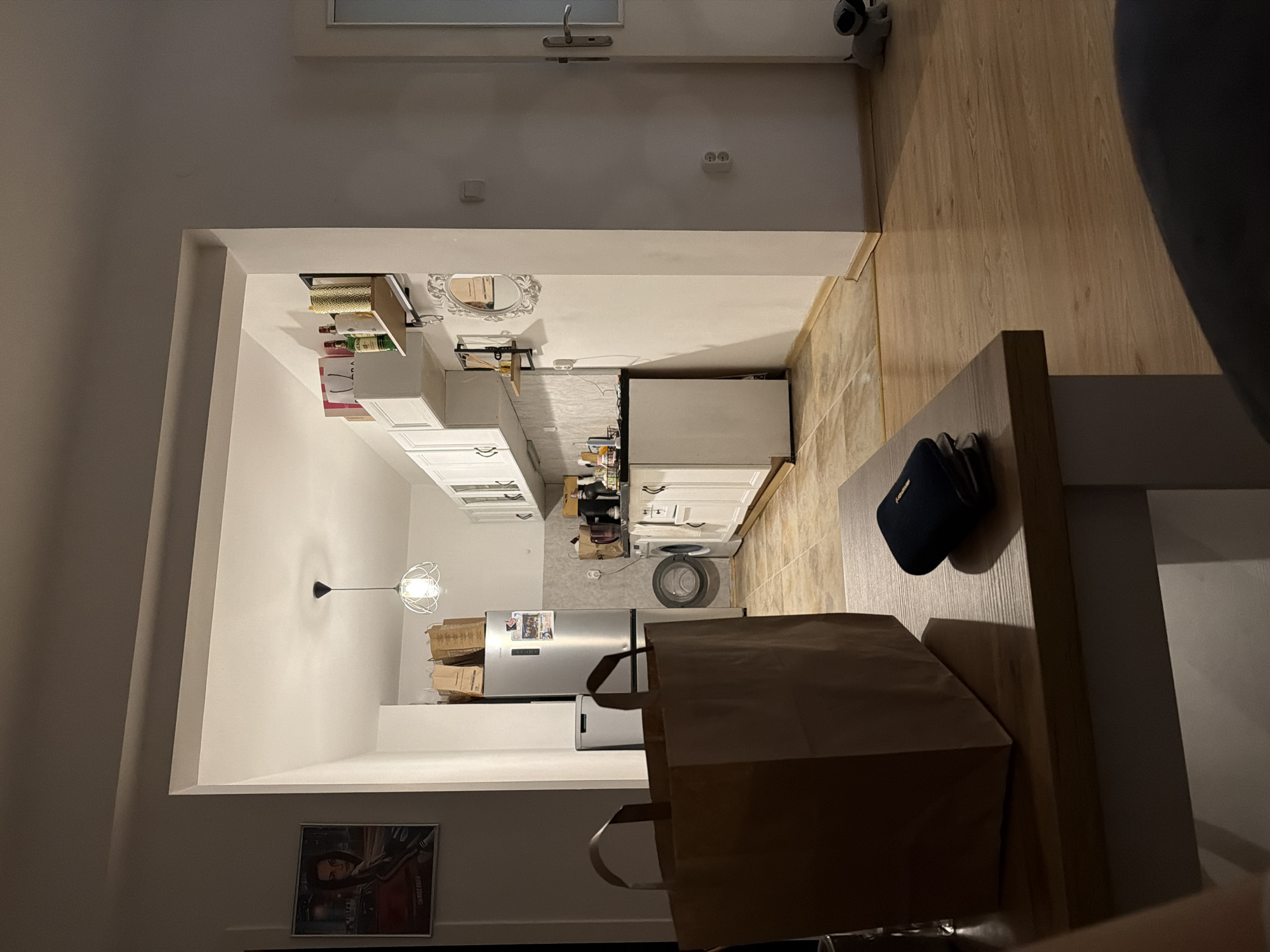 The image captures an interior view of a modern, open-plan kitchen and living/dining area. The perspective is from the living space looking into the kitchen.
In the foreground, a light wooden table or countertop is visible, on which a large brown paper bag with handles rests, along with a dark blue bag or speaker. To the left, on the wall, there is a framed picture depicting a person with dark hair and a jacket, with faint text "OLD SKOOL" visible. A modern-style light fixture hangs from the ceiling further into the room.
The kitchen area features white upper and lower cabinets and white countertops. A stainless steel refrigerator stands prominently, and next to it, a washing machine is visible under a countertop. Various items are placed on the shelves and countertops, including bottles of "JAMEISON" (Jameson whiskey) and "ABSOLUT" (Absolut vodka), along with other boxes and decorative items, such as a baroque-style mirror on the wall. The kitchen floor appears to be tiled, transitioning to light wooden flooring in the adjacent living area. Several cardboard boxes are stacked in various locations, suggesting recent activity like moving or storage.
The lighting is artificial, giving the scene an evening or night-time ambiance. No people are visible, and no specific activity is taking place. The space appears clean and functional. There are no direct visual cues within the image to indicate the specific location of Plovdiv, Bulgaria, provided in the context.