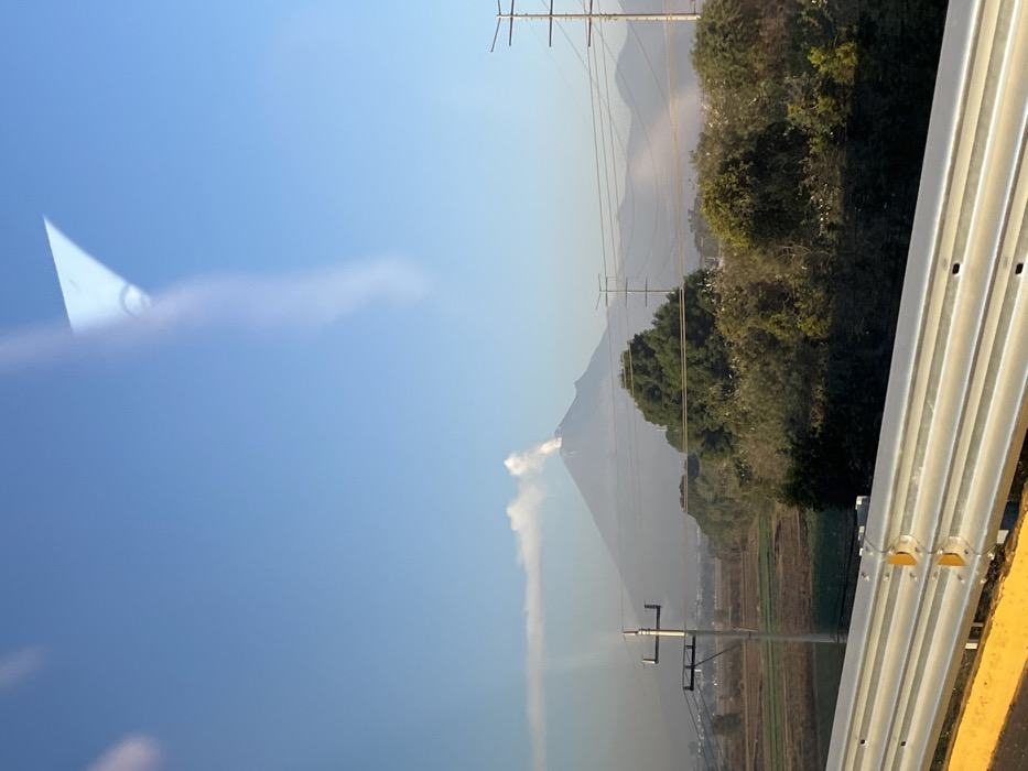 The image displays an outdoor landscape viewed through a window, evident by reflections across the upper sky and a vertical light streak on the left. In the immediate foreground, a metallic, multi-bar highway guardrail with a yellow base spans the bottom quarter of the frame. Beyond the guardrail, the midground features agricultural fields, alternating between tilled brown soil and green vegetation, extending from the left towards a line of dense green trees and shrubs that stretch across the frame. Multiple utility poles with power lines intersect the scene horizontally and vertically at various points. In the background, under a clear, bright blue sky, a large, conical stratovolcano, identified as Popocatépetl, dominates the central view. A prominent plume of white smoke or ash actively emanates from the volcano's summit, drifting horizontally to the left. Distant, hazy mountain ranges are visible to the right and behind the primary volcano. The vantage point is consistent with an area in or near Santa Ana Xalmimilulco, Mexico, looking towards the active volcano. No individuals or animals are visible.
