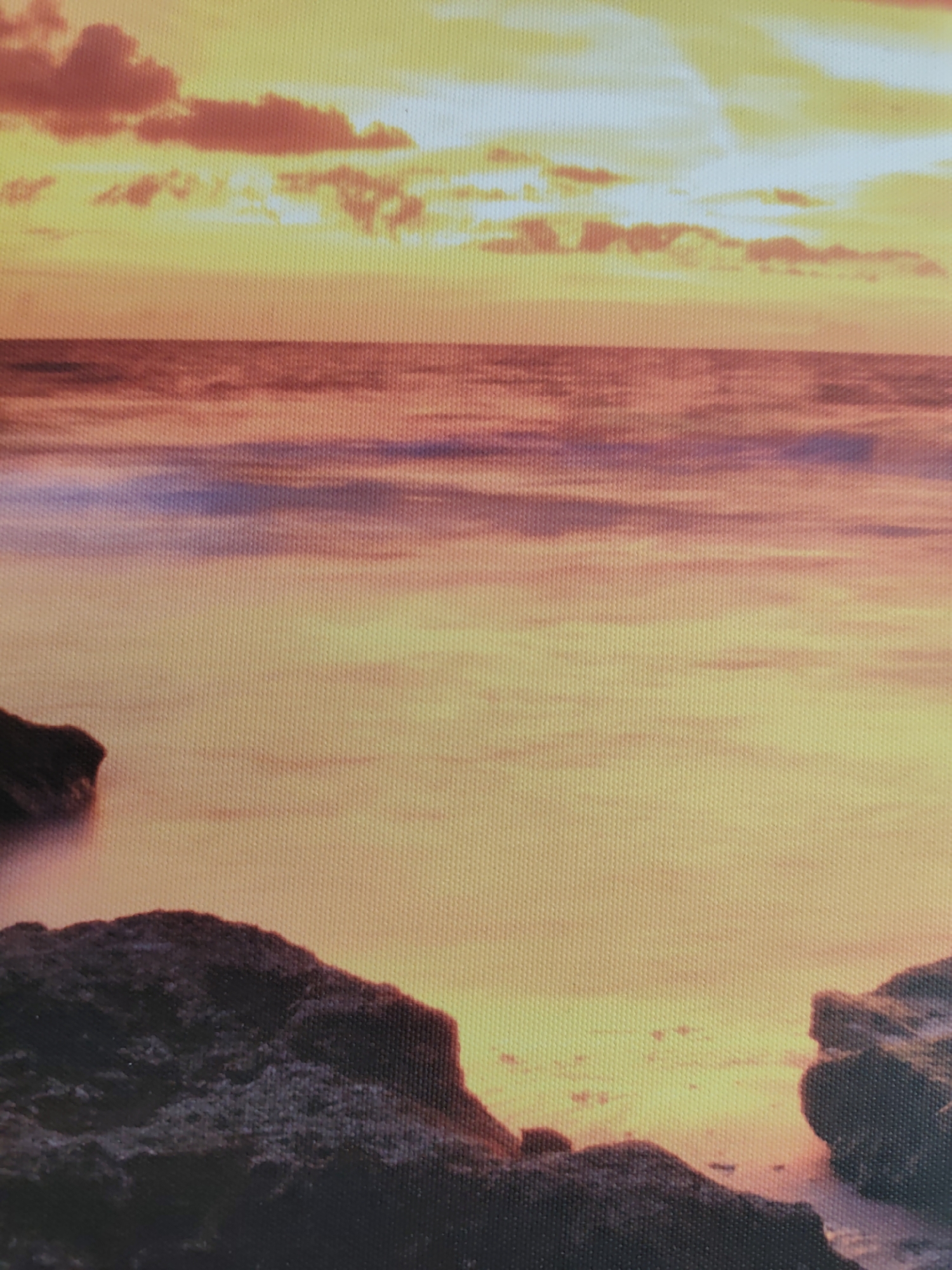 This image, a photographic print observed in the city of Pazardzhik, Bulgaria, depicts a long-exposure seascape during either sunrise or sunset. The scene is dominated by warm colors. The upper half features a gradient sky, transitioning from bright yellow and orange at the horizon to deeper reds and oranges higher up, with visible, scattered clouds. The horizon line is flat and distinct. Below the horizon, a body of water, likely an ocean or sea, displays a smooth, painterly surface due to the long exposure, reflecting the sky's hues in shades of orange, red, and subtle purple-blue where waves are blurred. In the foreground, dark, rugged rock formations anchor the bottom left and right corners, appearing as silhouettes against the brighter water and sky. There are no people, animals, or specific objects other than the natural elements depicted. The entire image exhibits a visible grid-like texture, indicative of printing on a woven material such as canvas.