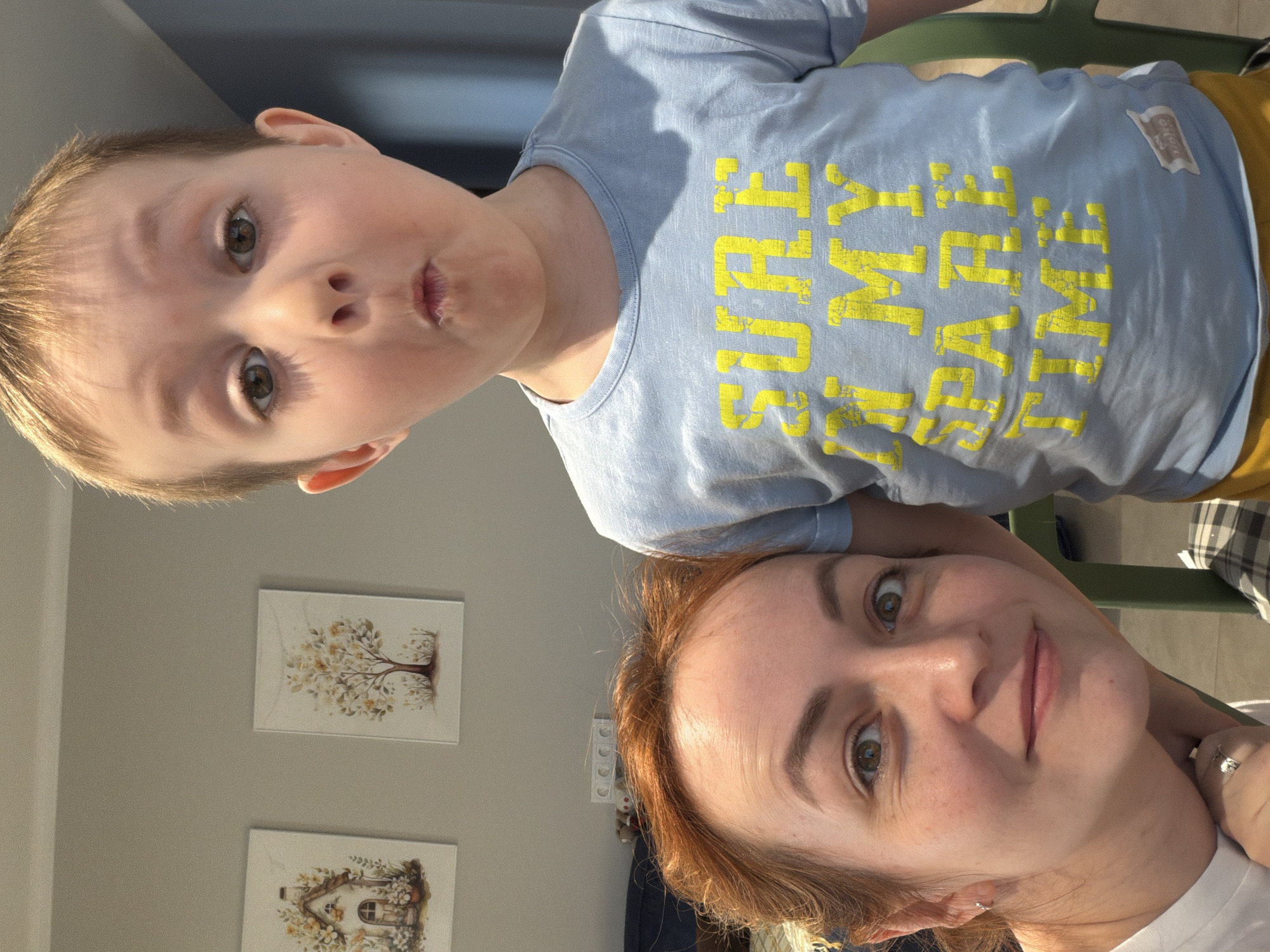 A woman and a young boy are in a selfie photograph. The woman has fair skin, green eyes, and auburn hair pulled back. The boy has fair skin, light blonde hair, and brown eyes. He is wearing a light blue t-shirt with yellow writing, and his mouth is puckered. The background includes a grey wall with two framed pictures, an electrical outlet, and part of a green chair. The location is Chaikovsky, Russia.