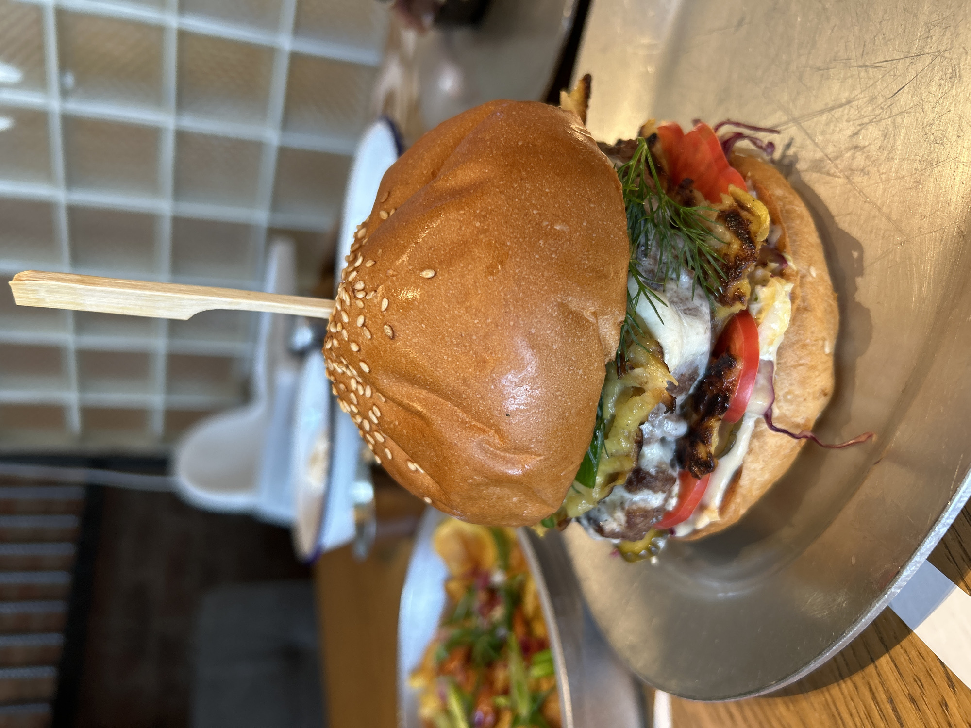 A close-up, eye-level shot showcases a large, gourmet burger served on a metallic plate. The burger is constructed with a golden-brown sesame seed bun, a thick meat patty, melted cheese, fresh lettuce, tomato slices, pickles, and a sprig of dill, all secured with a wooden skewer. To the left, out of focus, a silver bowl filled with crispy fries adorned with green onions and red garnishes is visible. The background is blurred, revealing a rustic interior with brick walls, frosted glass partitions, and wooden tables, suggesting a restaurant setting. The lighting is natural and soft, highlighting the textures and colors of the food. The image focuses on the burger, with the surrounding elements providing context.